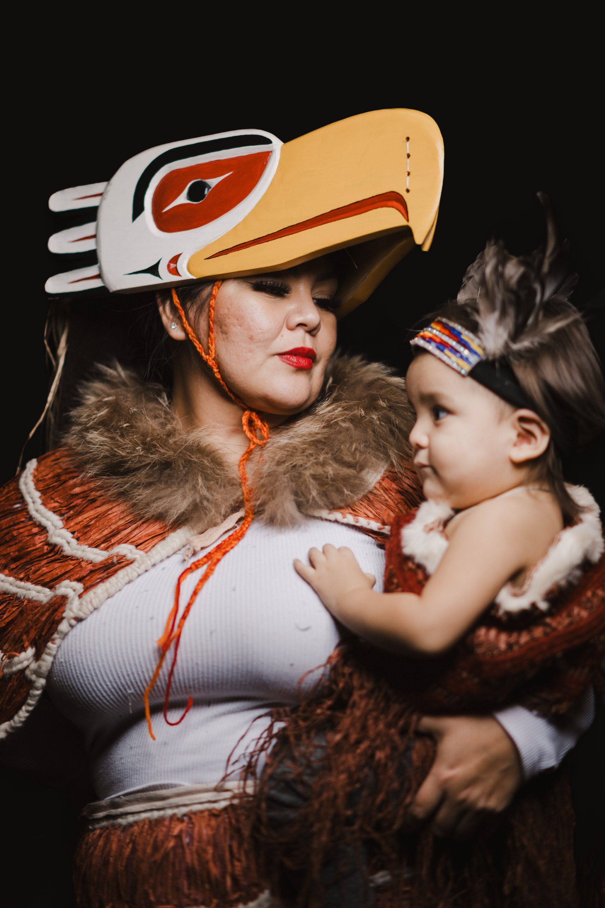 Woman and child in Coast Salish regalia with carved eagle headdress and fur wraps, embracing against black backdrop