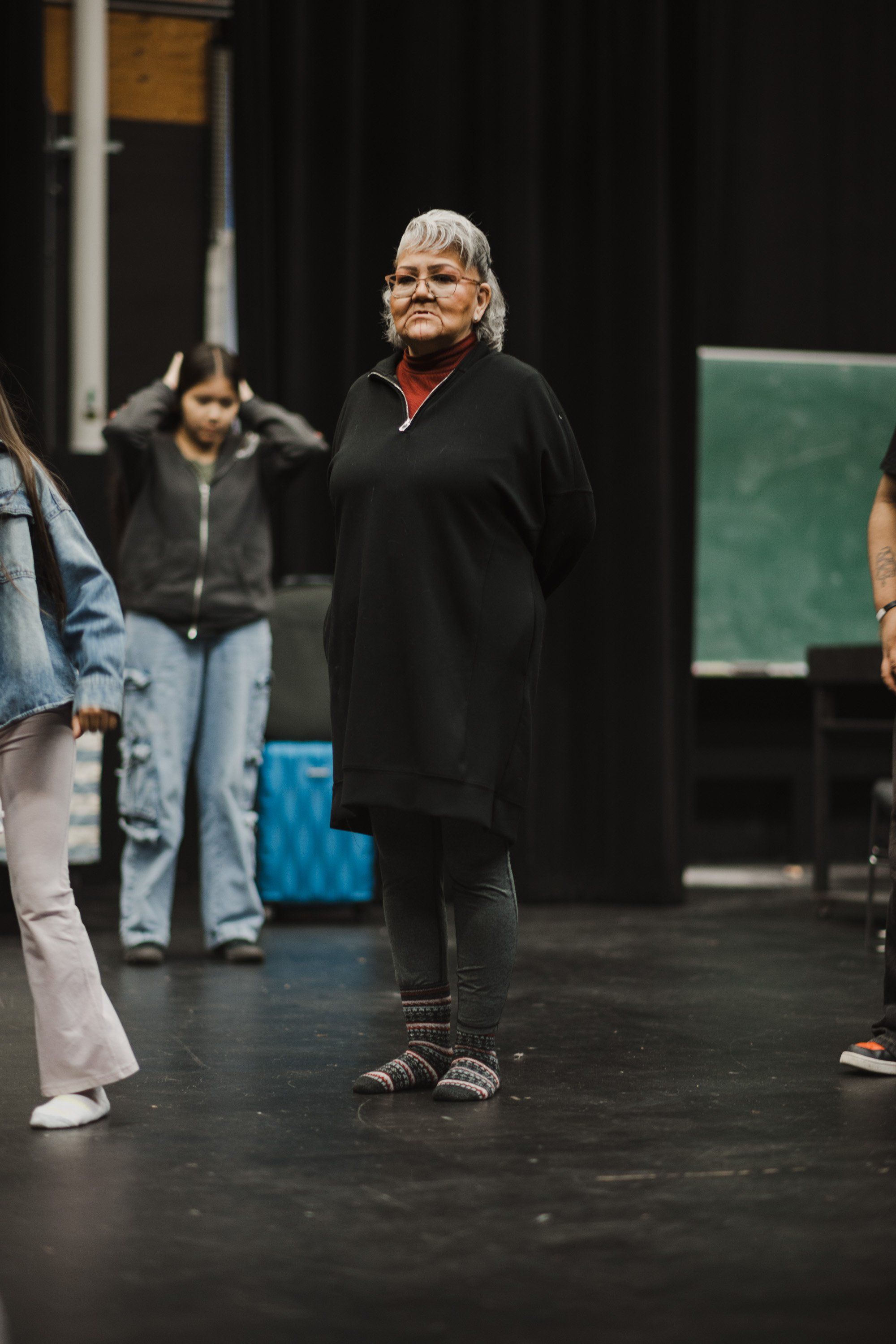Older woman with white hair and glasses in black sweater stands on dark stage, smiling warmly at audience during Coast Salish cultural performance