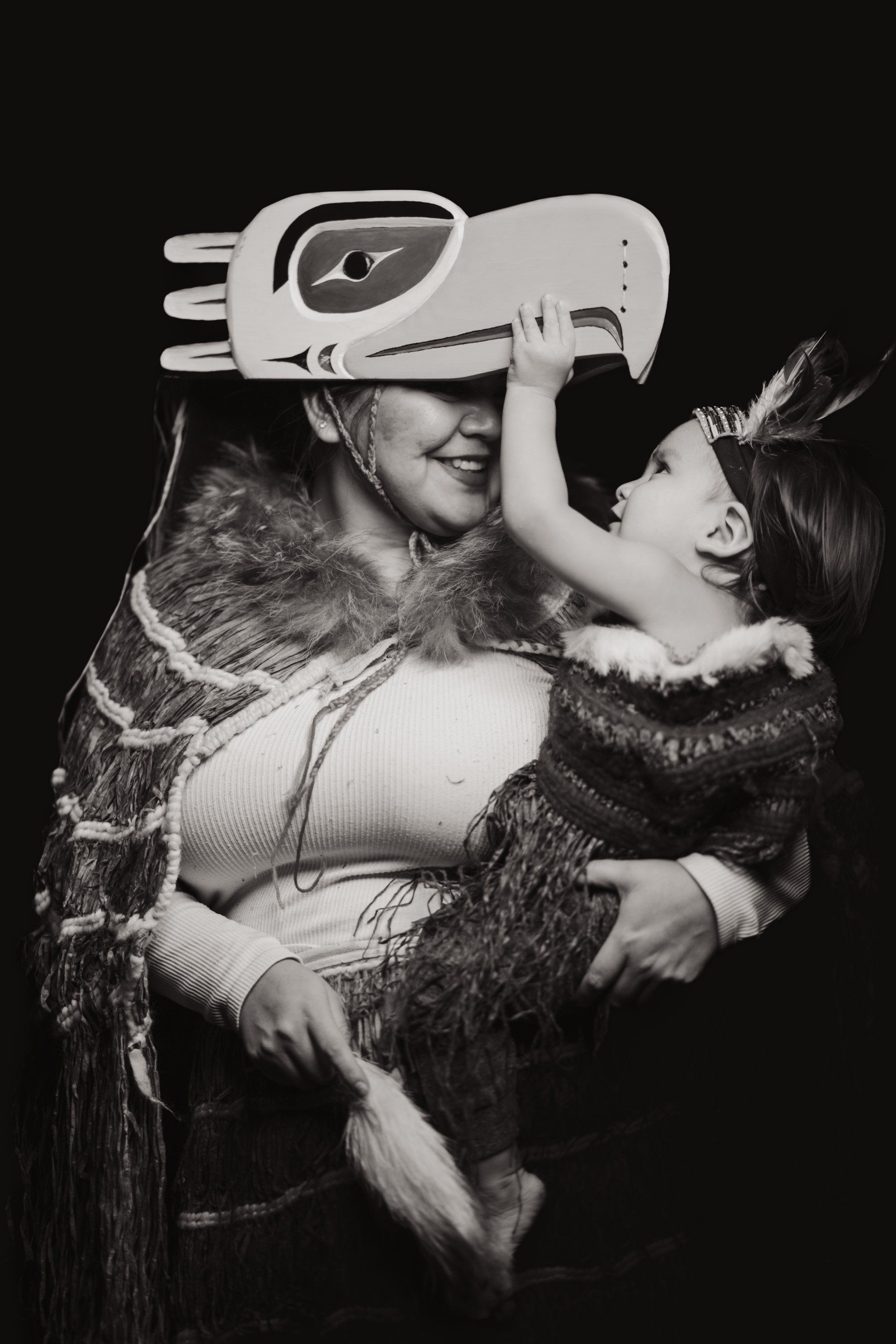 Man in traditional regalia holding child wearing wolf mask against black background, intimate family portrait in dramatic studio lighting