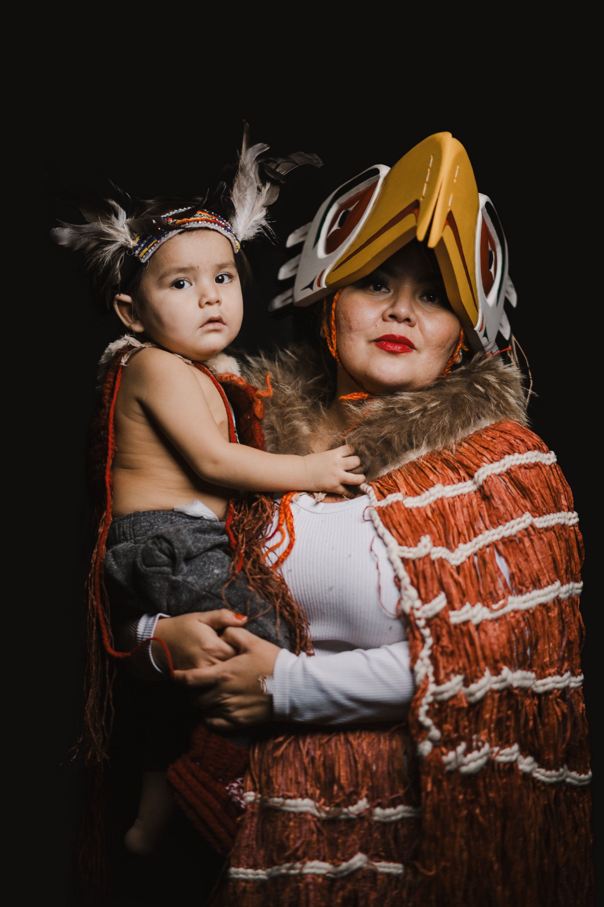 Woman in ochre hat and rust-patterned regalia holds toddler wearing feathered headband against dark background