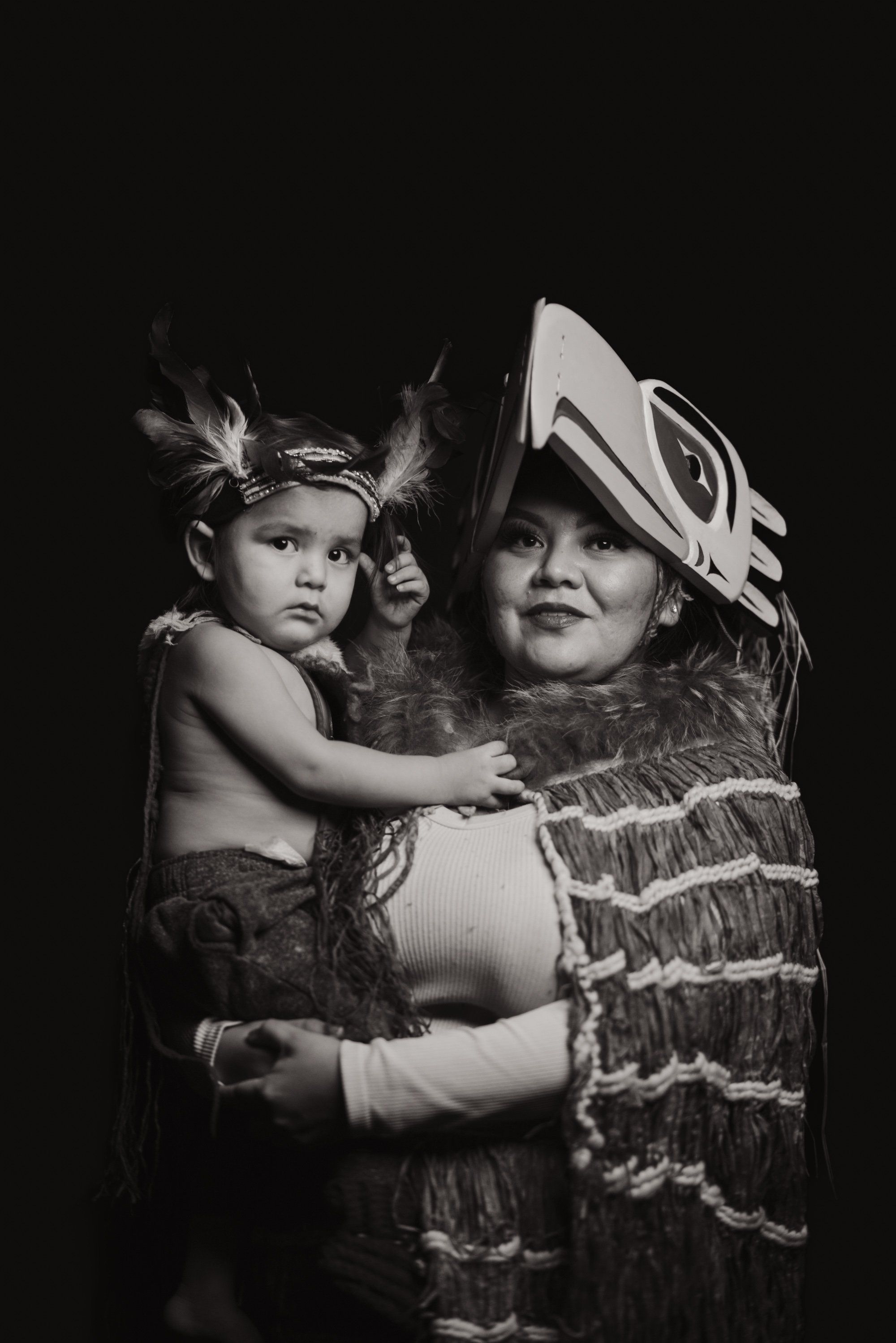 Two children in traditional Coast Salish regalia embrace against black, their carved masks and woven blankets illuminated in striking monochrome
