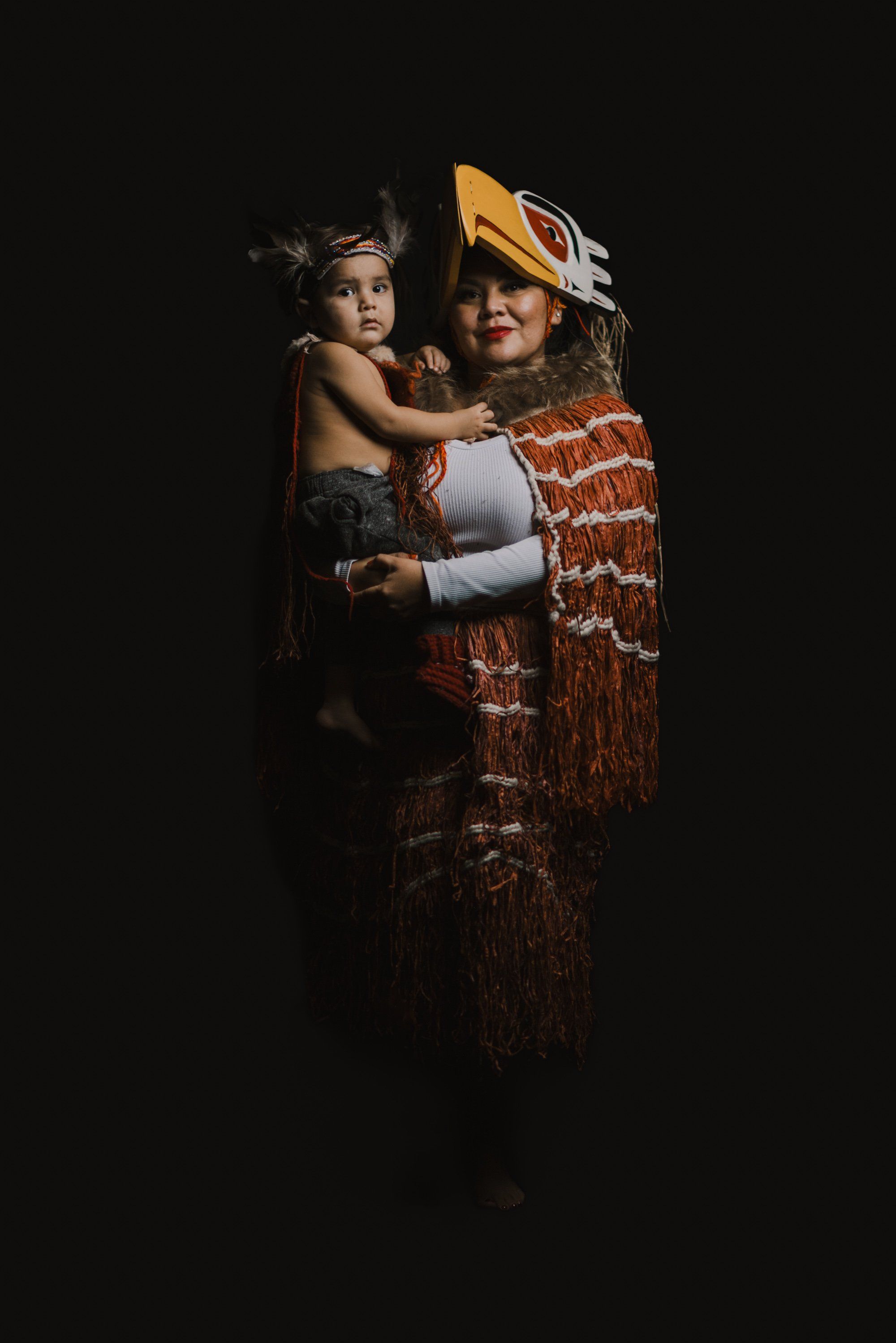 Two figures in traditional regalia embrace against black, with gold button blanket, cedar hat, and white gloves creating striking contrast
