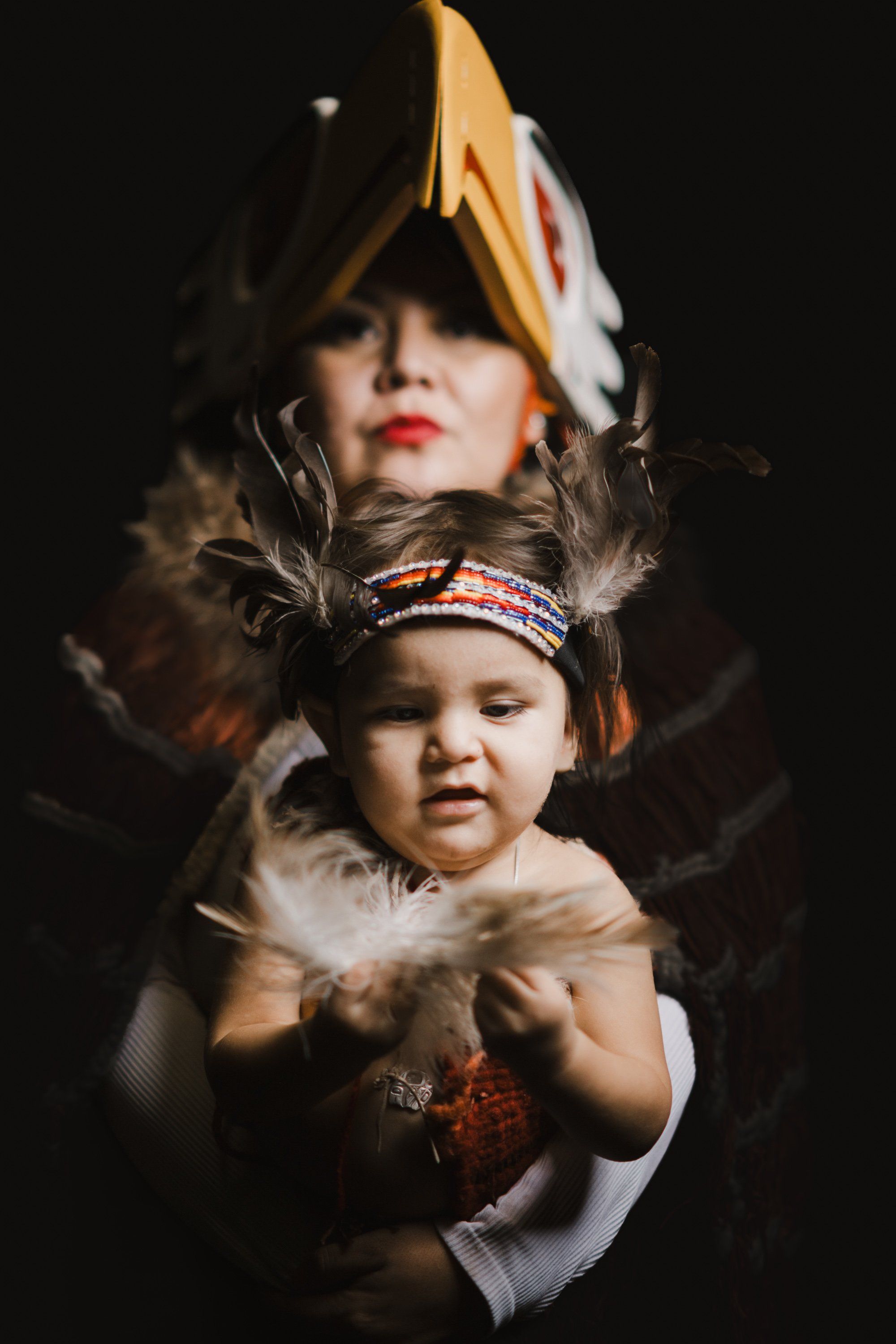 Two children in traditional Coast Salish regalia pose against a dark background, the older child wearing a carved wooden hat and the younger in beaded headba…