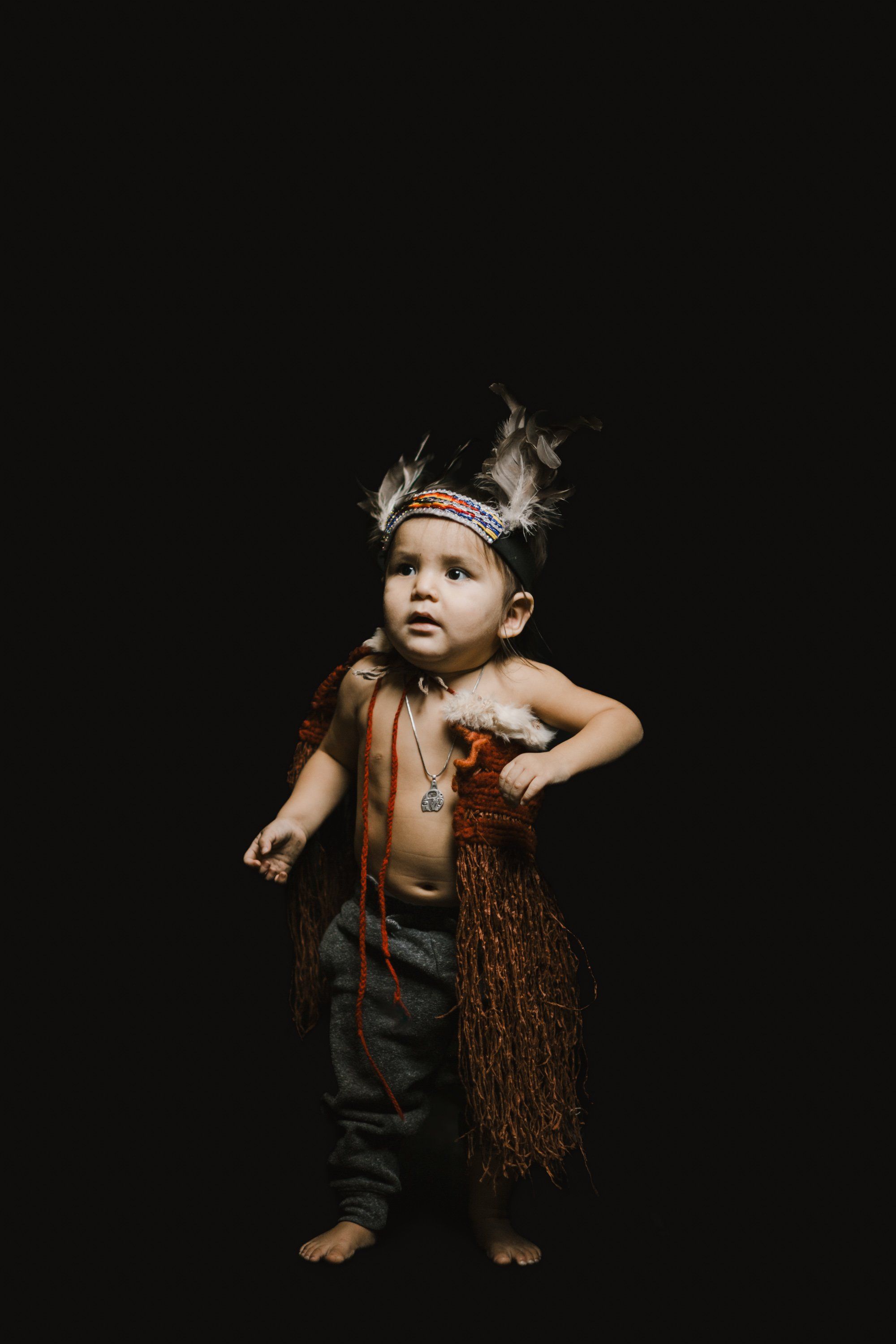 Toddler in traditional regalia with feathered headpiece and woven cedar skirt stands against black background, gazing upward