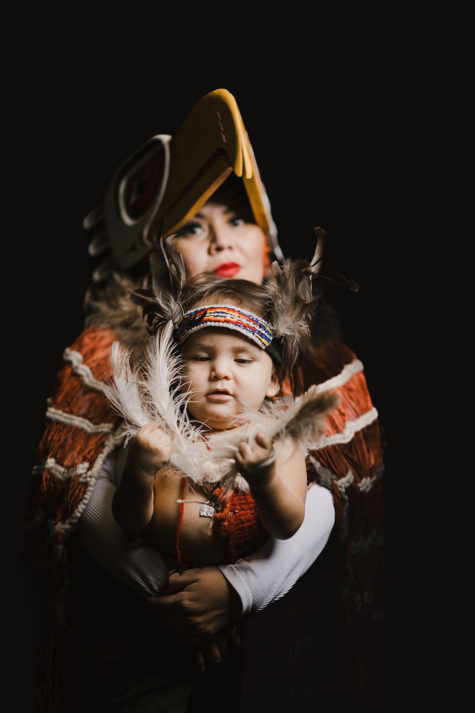 An adult and child wear traditional regalia with feathered collars and golden headdresses against a black background, their faces painted and positioned clos…