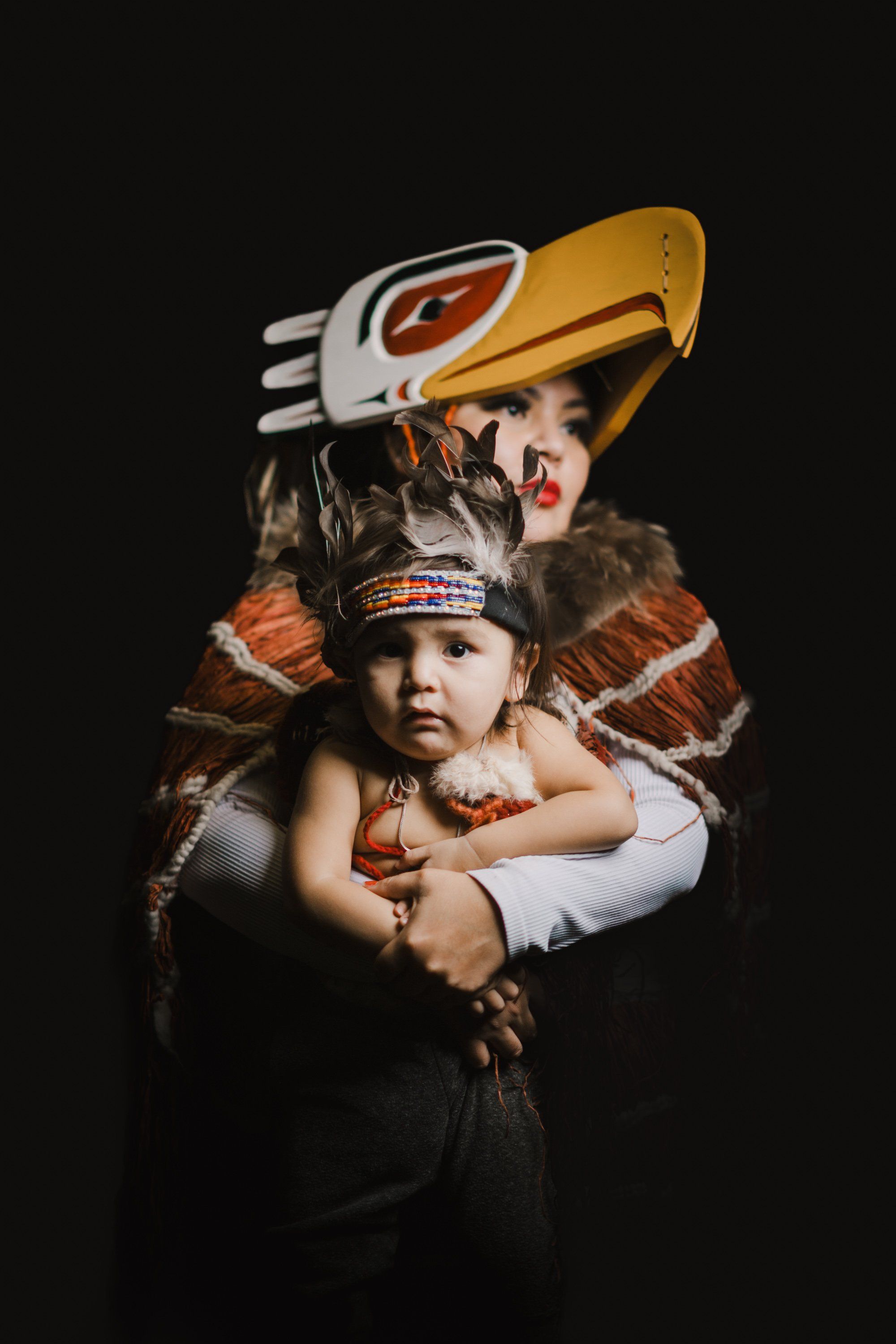 Child wearing traditional Coast Salish regalia with formline art designs, held by an adult in ceremonial button blanket and carved wooden mask against black…