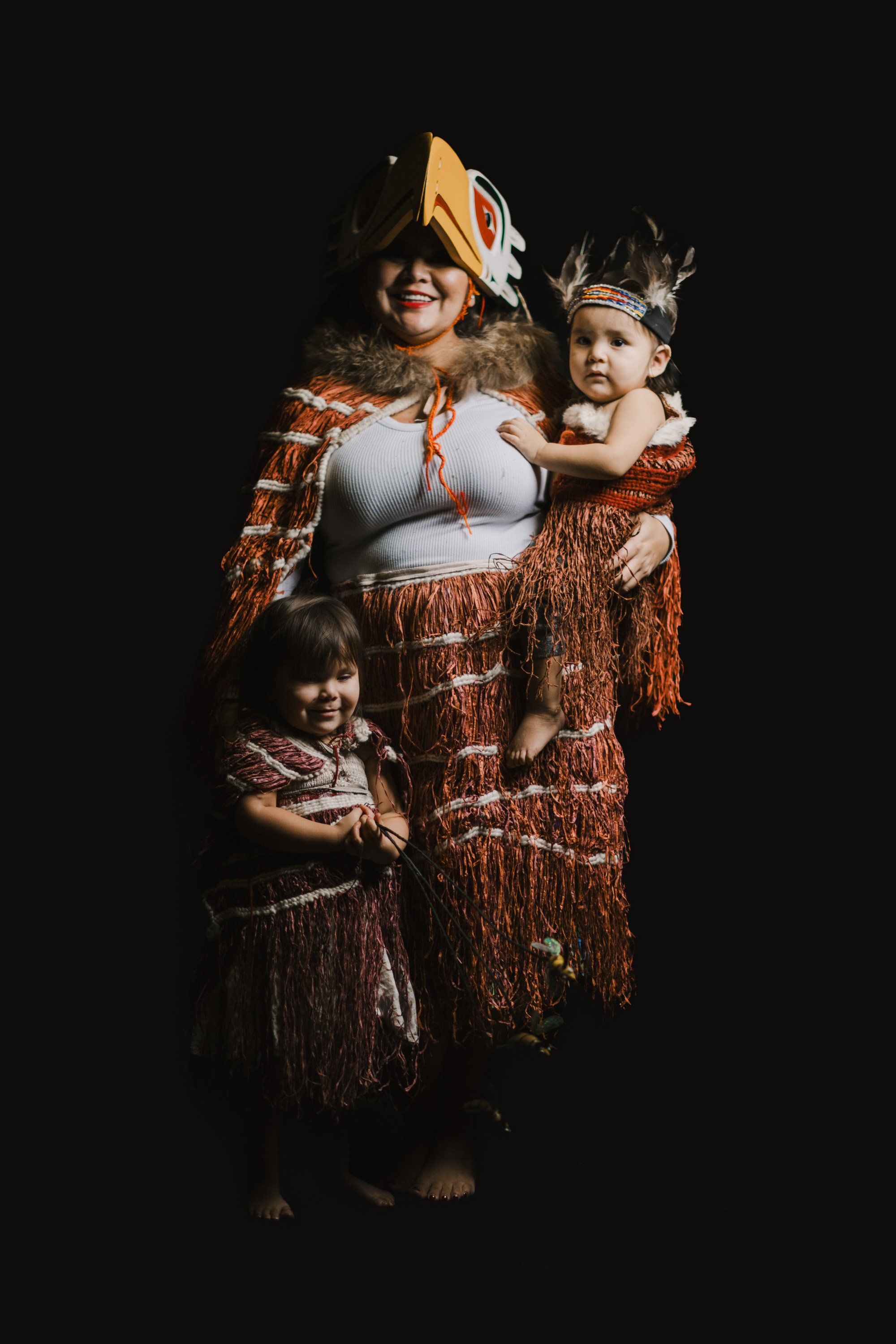 Woman and two children in copper-fringed regalia and masks pose against black background, celebrating Coast Salish heritage