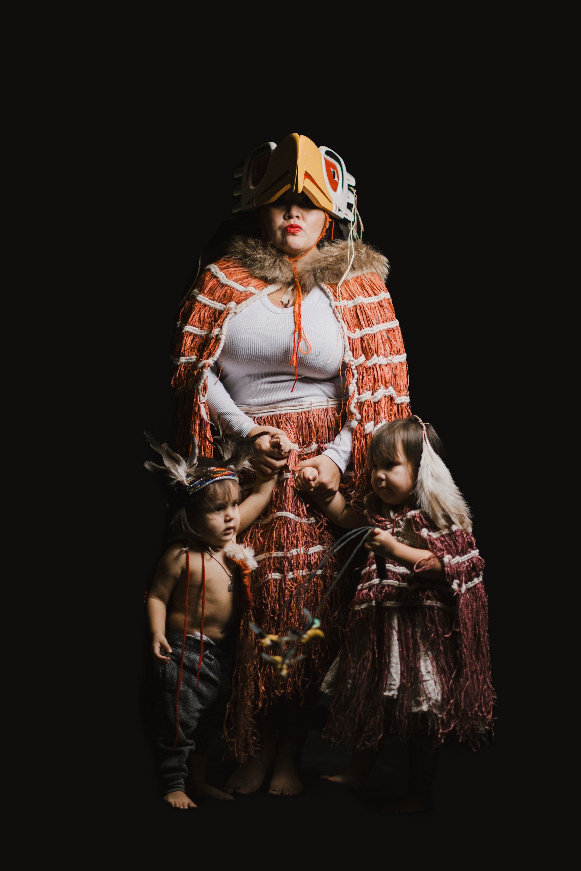 Woman in traditional Coast Salish regalia with orange and red woven blanket stands with two children against black background, wearing goggles and ceremonial…