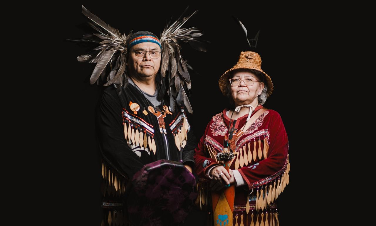 Man wearing glasses and feathered headdress with beaded regalia turns in profile against black background