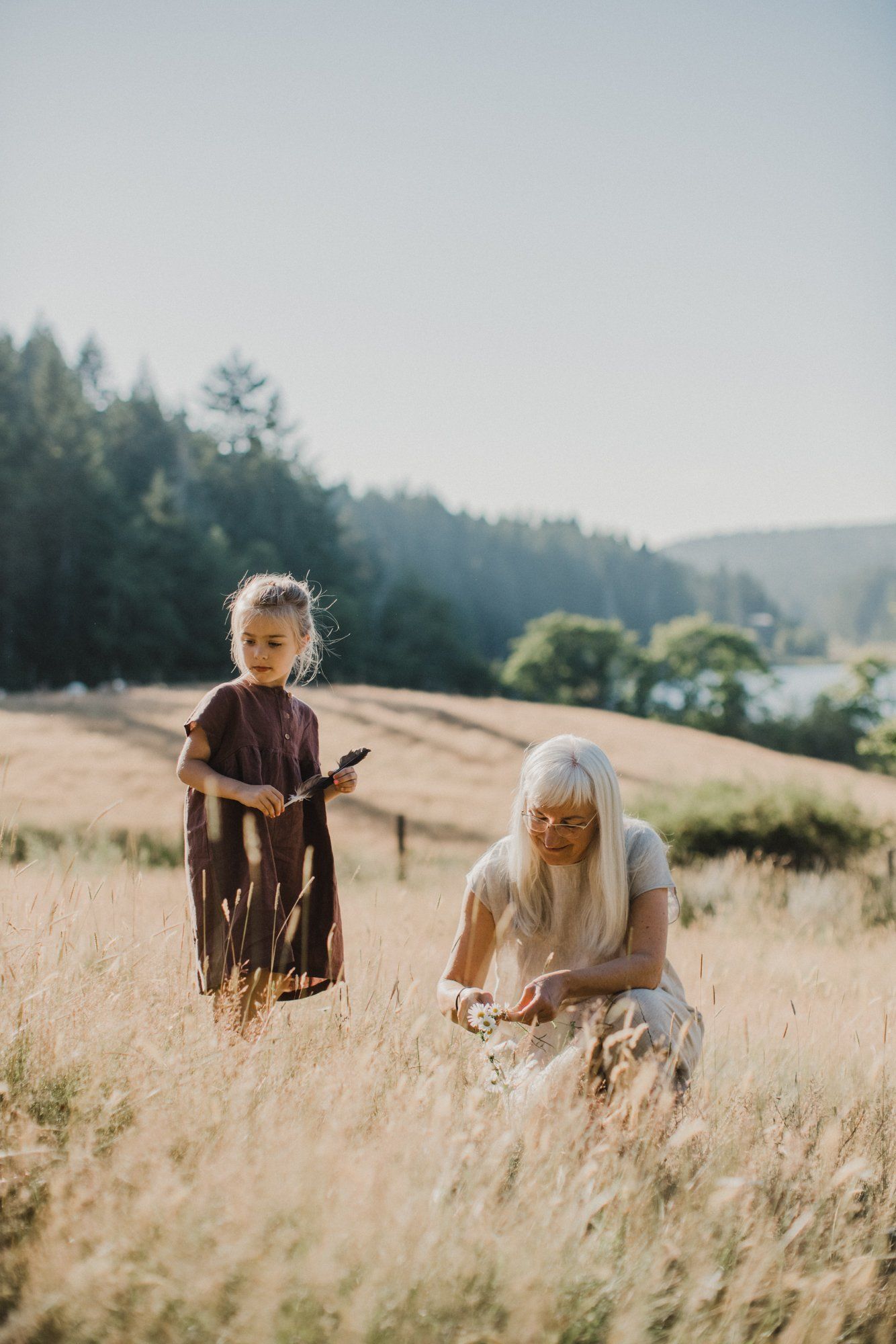 Two figures in earthy tones stand in a golden grain field with forested hills under soft, hazy light