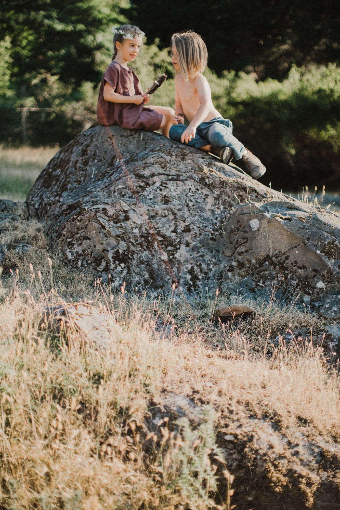 Two children sit facing each other atop a large boulder in golden afternoon light, surrounded by dry grass and evergreen trees
