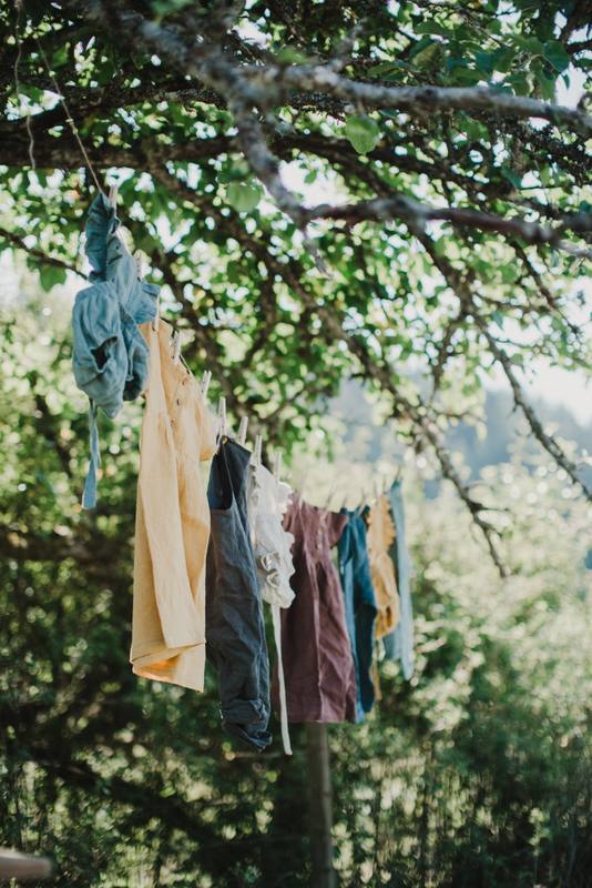 Handmade garments in cream, black, burgundy, and teal hang from a tree branch, dappled in golden sunlight and green foliage