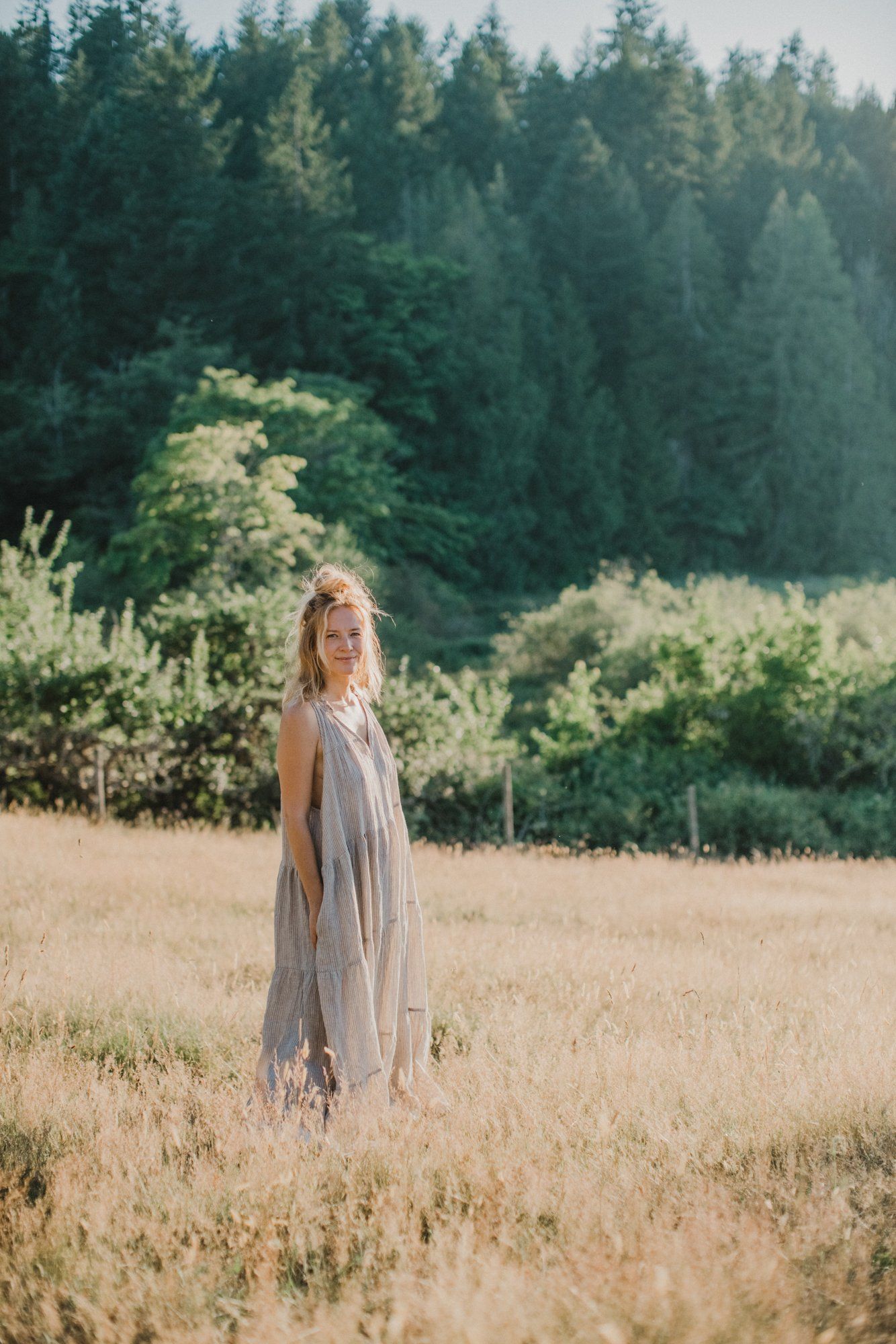 Woman in flowing gray dress standing in golden field with evergreen forest and soft afternoon light behind her