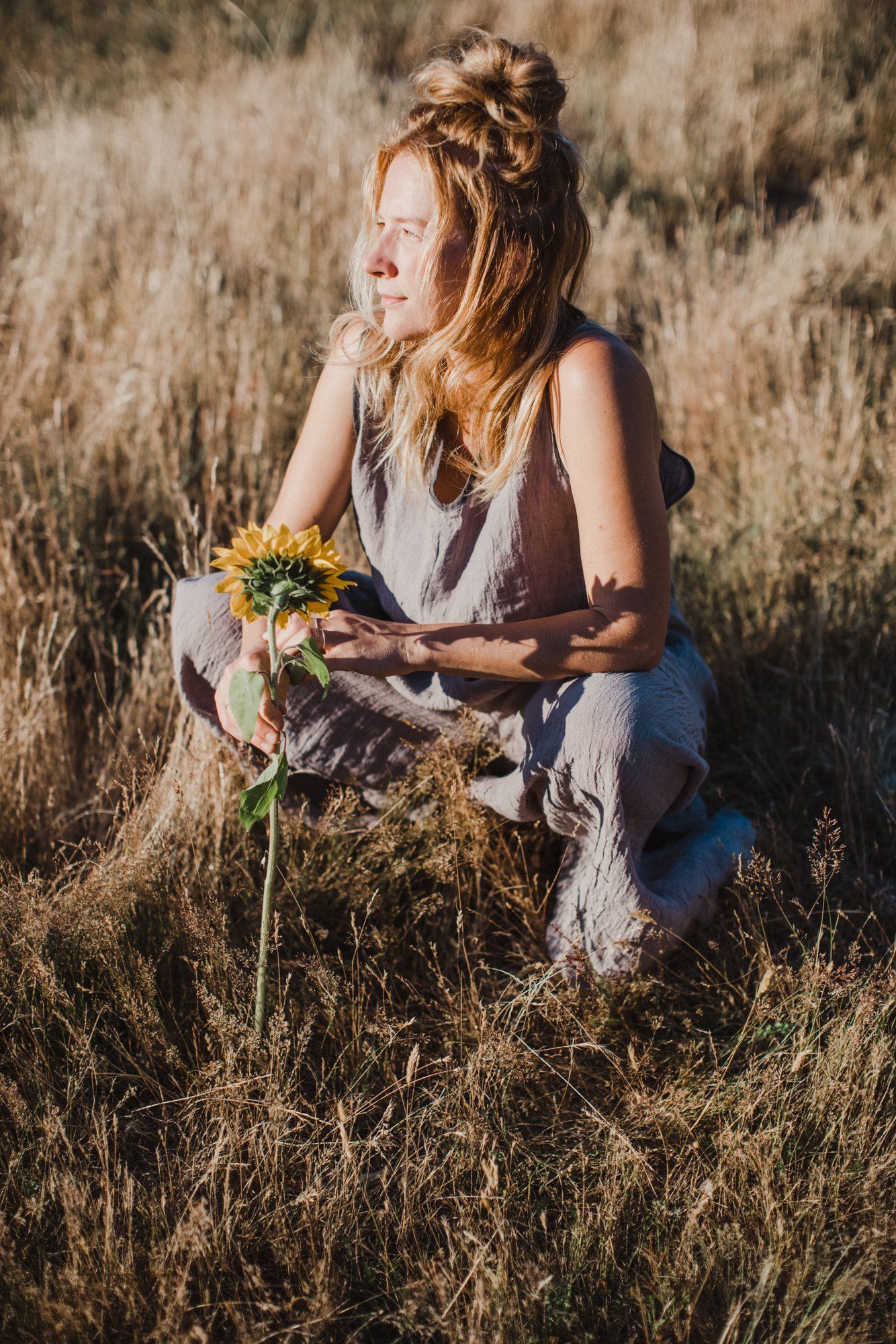 Redheaded woman in linen vest crouches in golden dry grass holding yellow wildflowers, gazing to the side in warm afternoon light