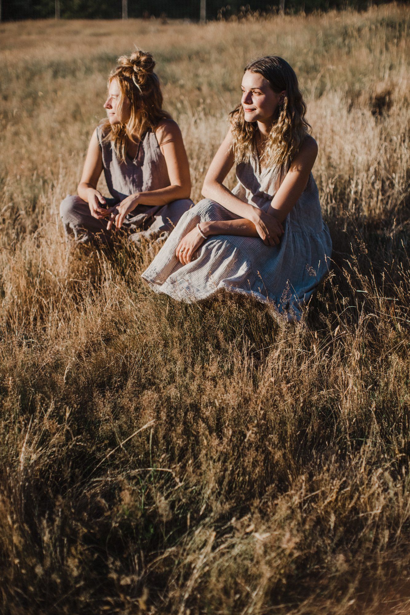 Two young women in light linen dresses sit together in a golden, dry meadow bathed in warm afternoon sunlight
