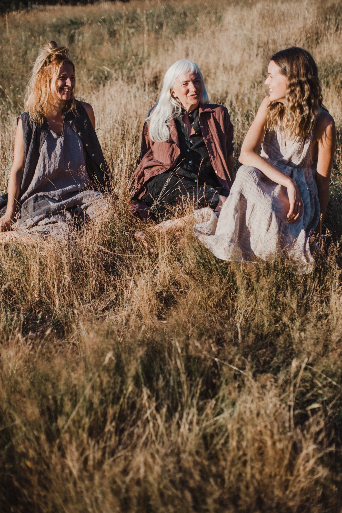 Three women sit together in golden dry grass at golden hour, wearing layered boho clothing in earth tones and burgundy