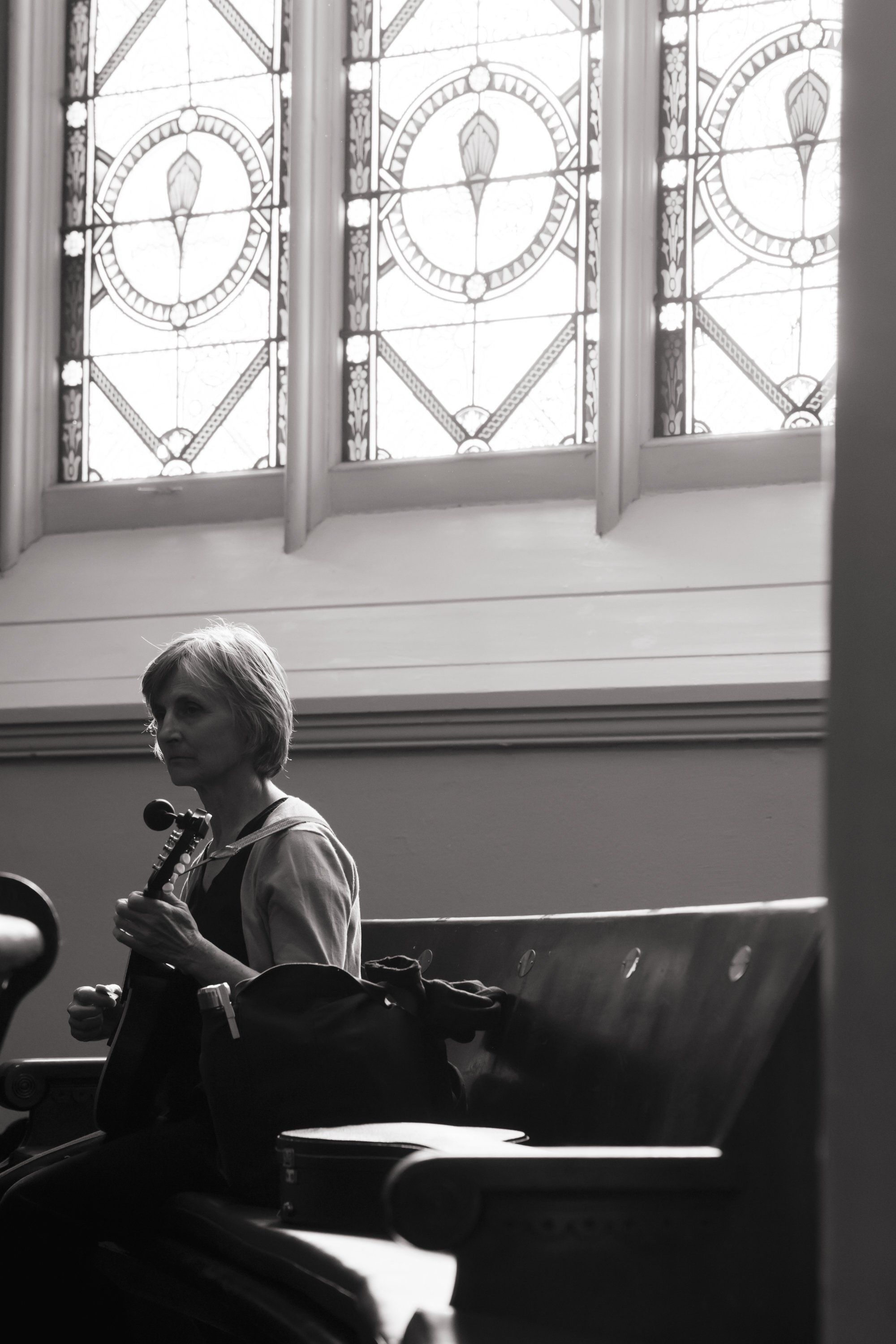 Musician seated on dark leather bench beneath ornate stained-glass windows, holding violin in contemplative pose