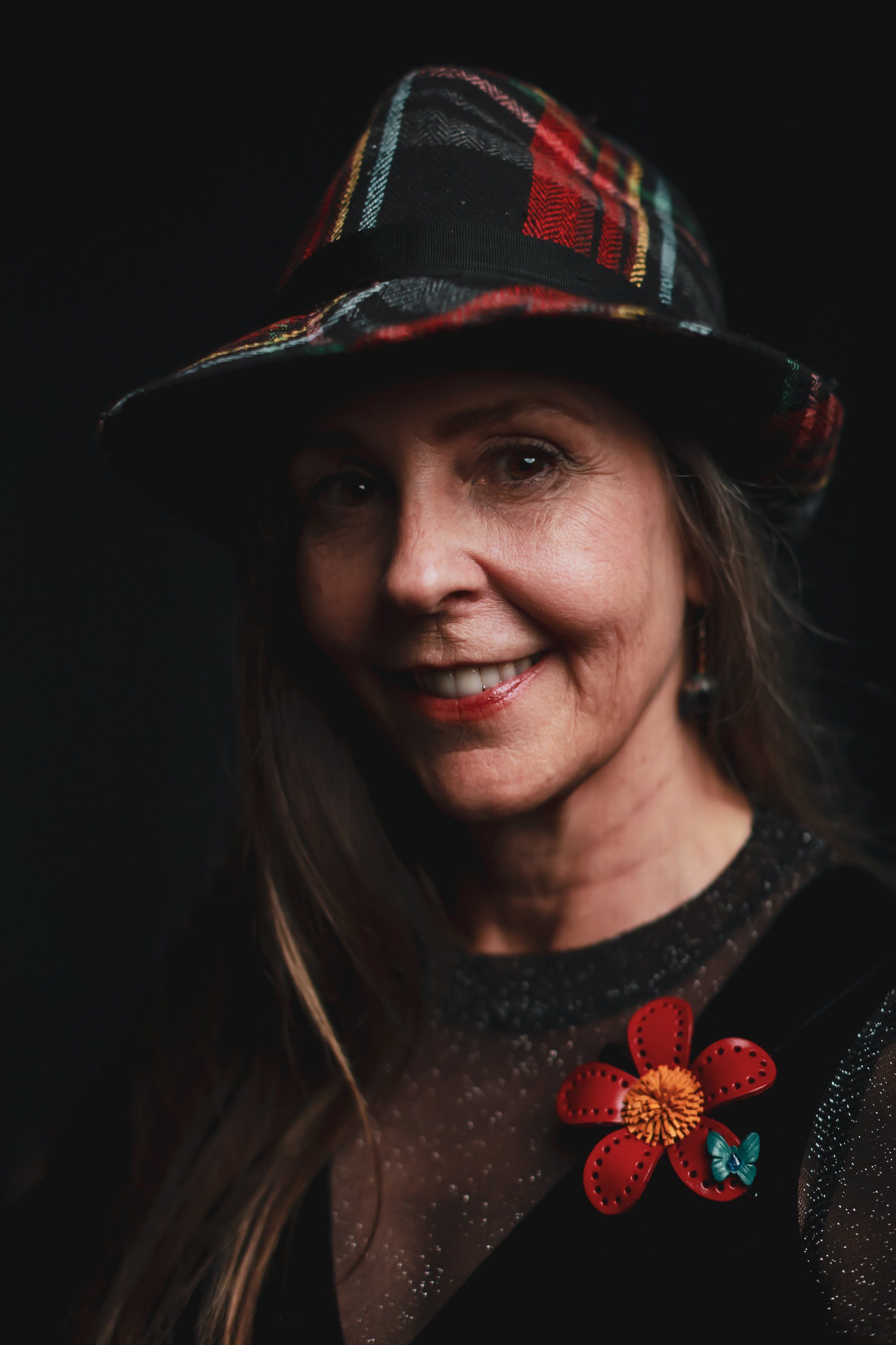 Smiling woman wearing a red and black plaid hat and sparkly dark sweater with a red floral brooch, photographed against a black background