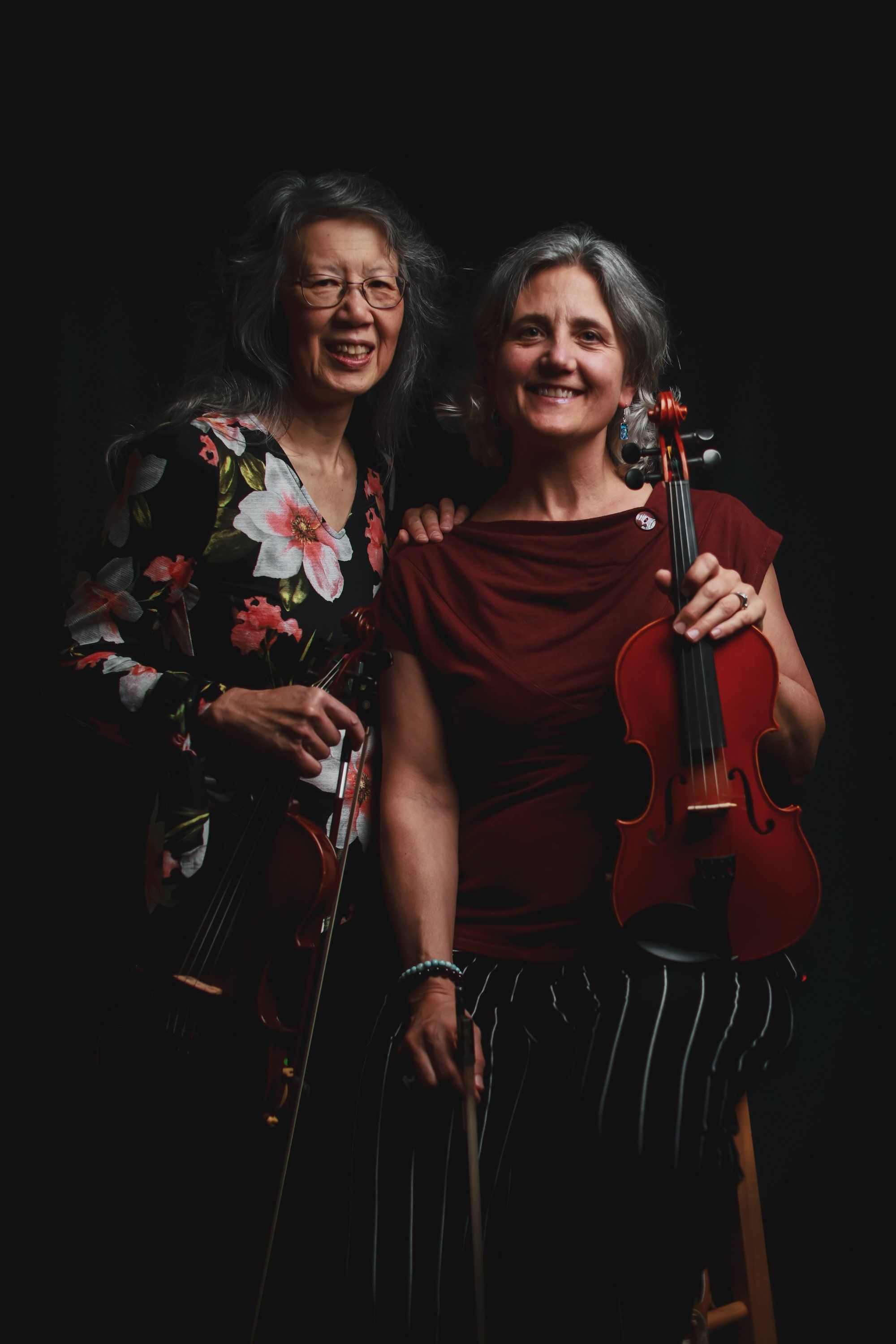 Two musicians with violins embrace warmly against a dark background, wearing colorful floral and burgundy clothing, smiling at the camera