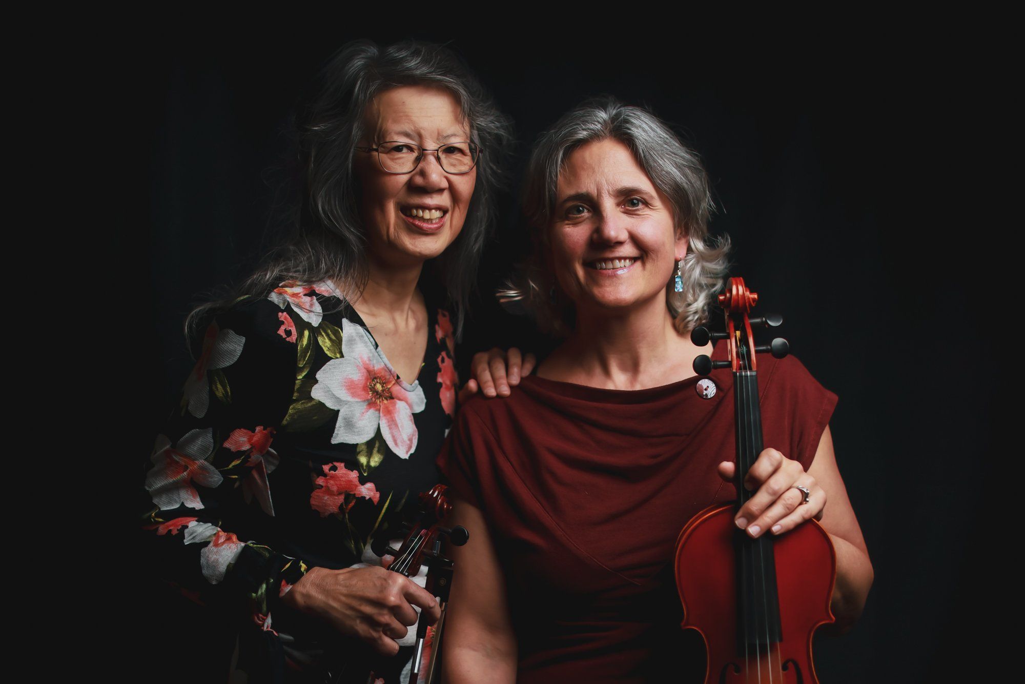 Two smiling women pose together against a black background, one wearing a floral print jacket and the other holding a violin in a burgundy top