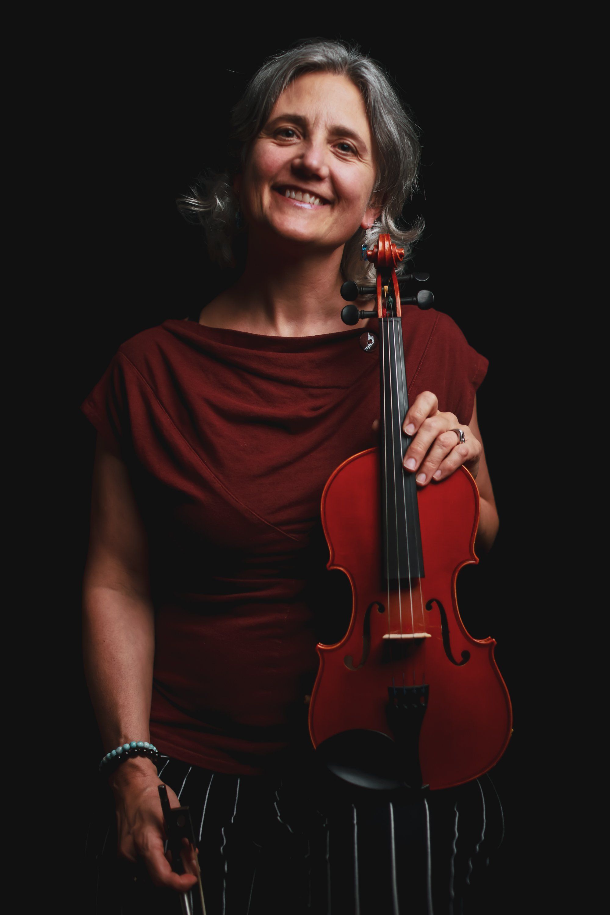 Smiling musician in burgundy shirt holds a red violin against black background, warmly lit with natural expression