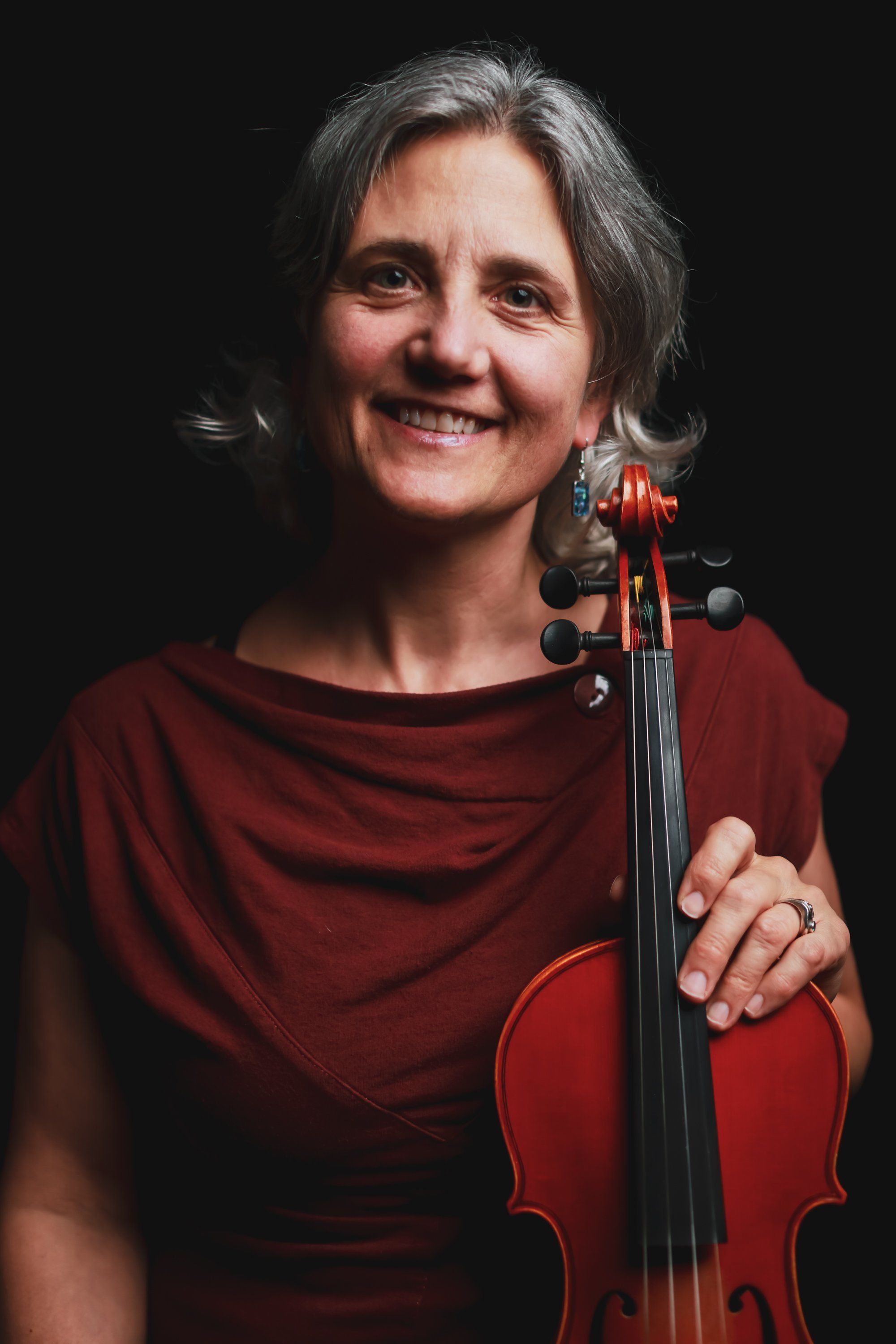 Smiling musician with gray hair holds a red viola against a black background, wearing a burgundy shirt