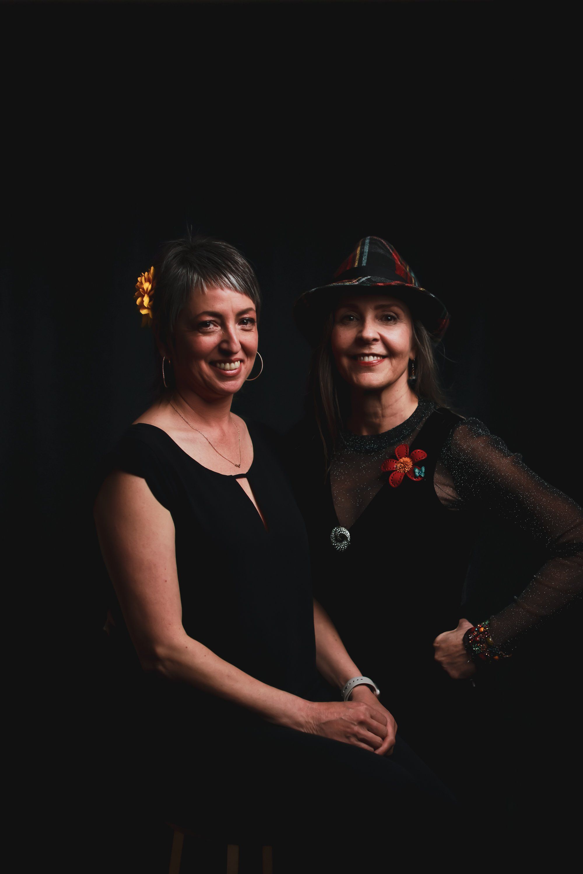 Two women smiling warmly against a black background, one in a simple black sleeveless top, the other wearing a black hat with red accents and sheer overlay
