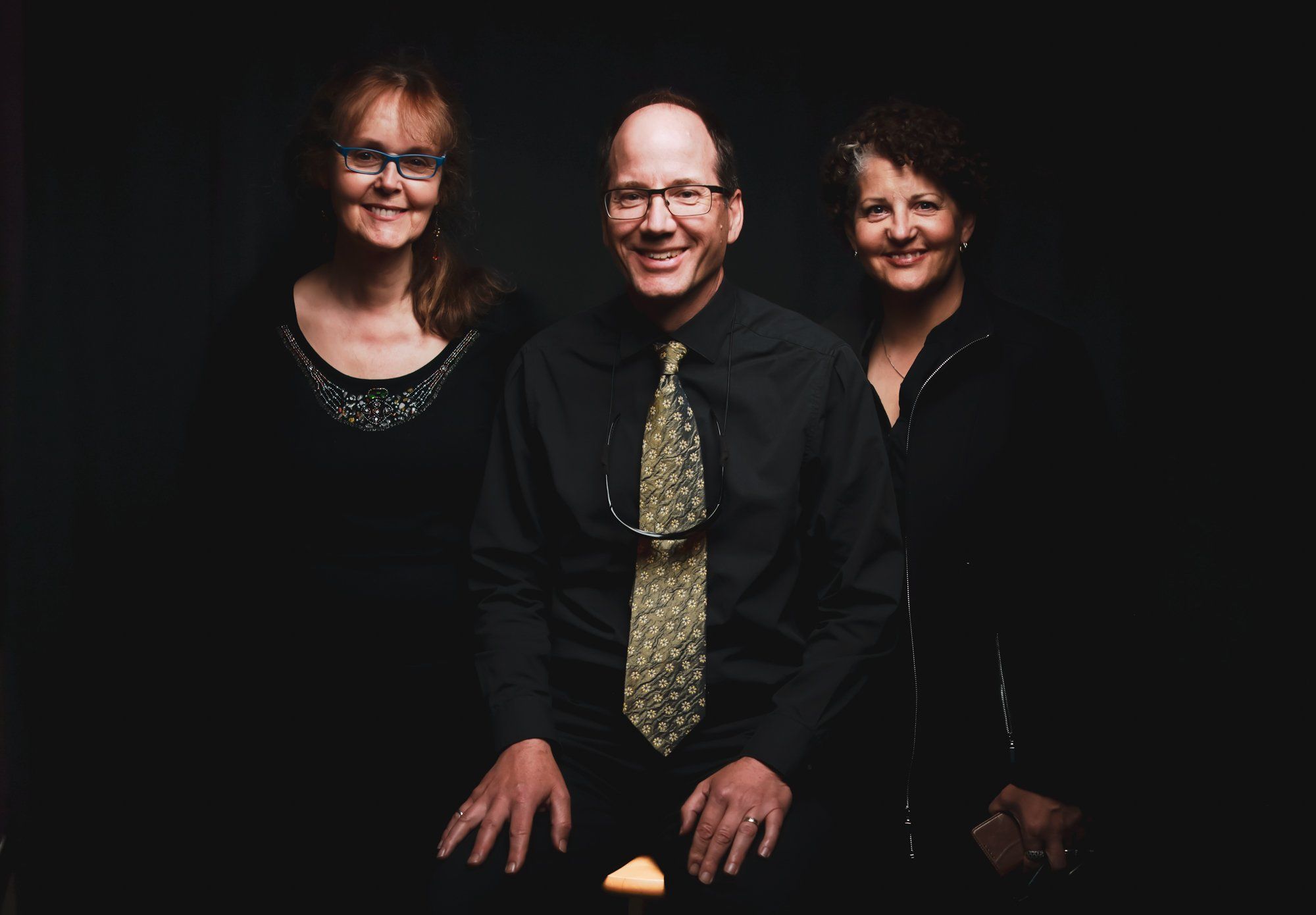 Three musicians smile warmly against a dark background, wearing formal attire with subtle lighting that highlights their faces and creates an intimate, profe…