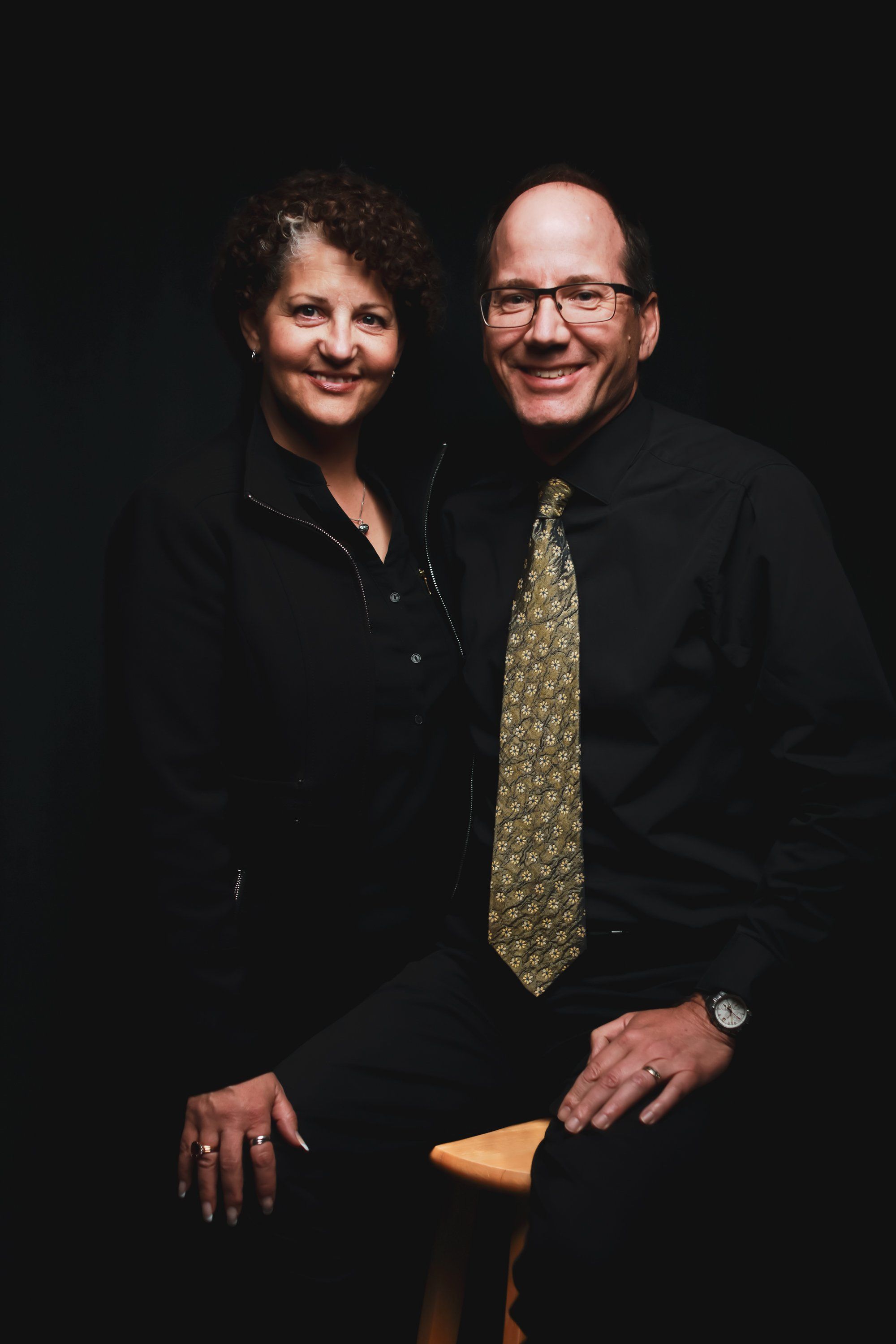 Two smiling adults in formal black clothing pose together against a dark background, warm studio lighting illuminating their faces and a gold-patterned tie