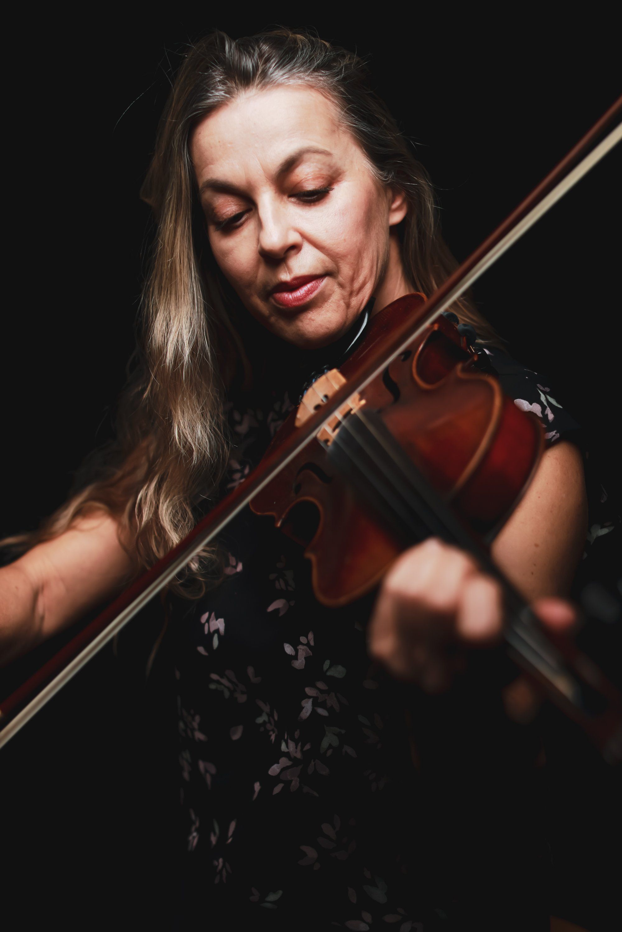 Violinist with eyes closed plays expressively against black backdrop, long hair catching warm light, instrument angled dynamically across frame