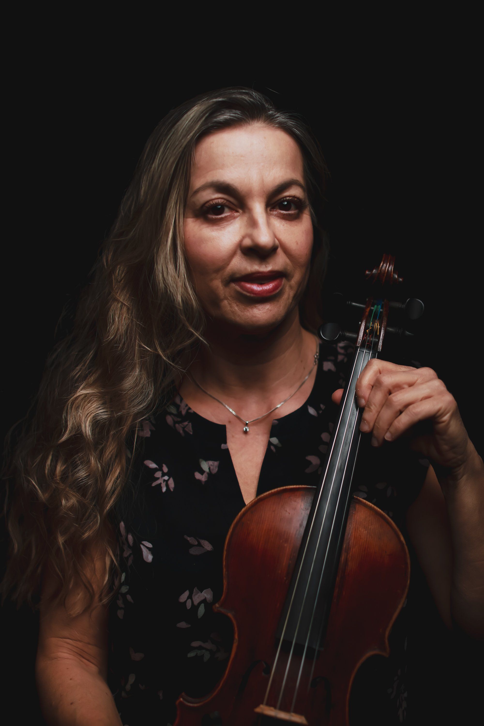 Violinist with long dark hair holds her instrument against a black background, wearing a dark floral top and warm smile