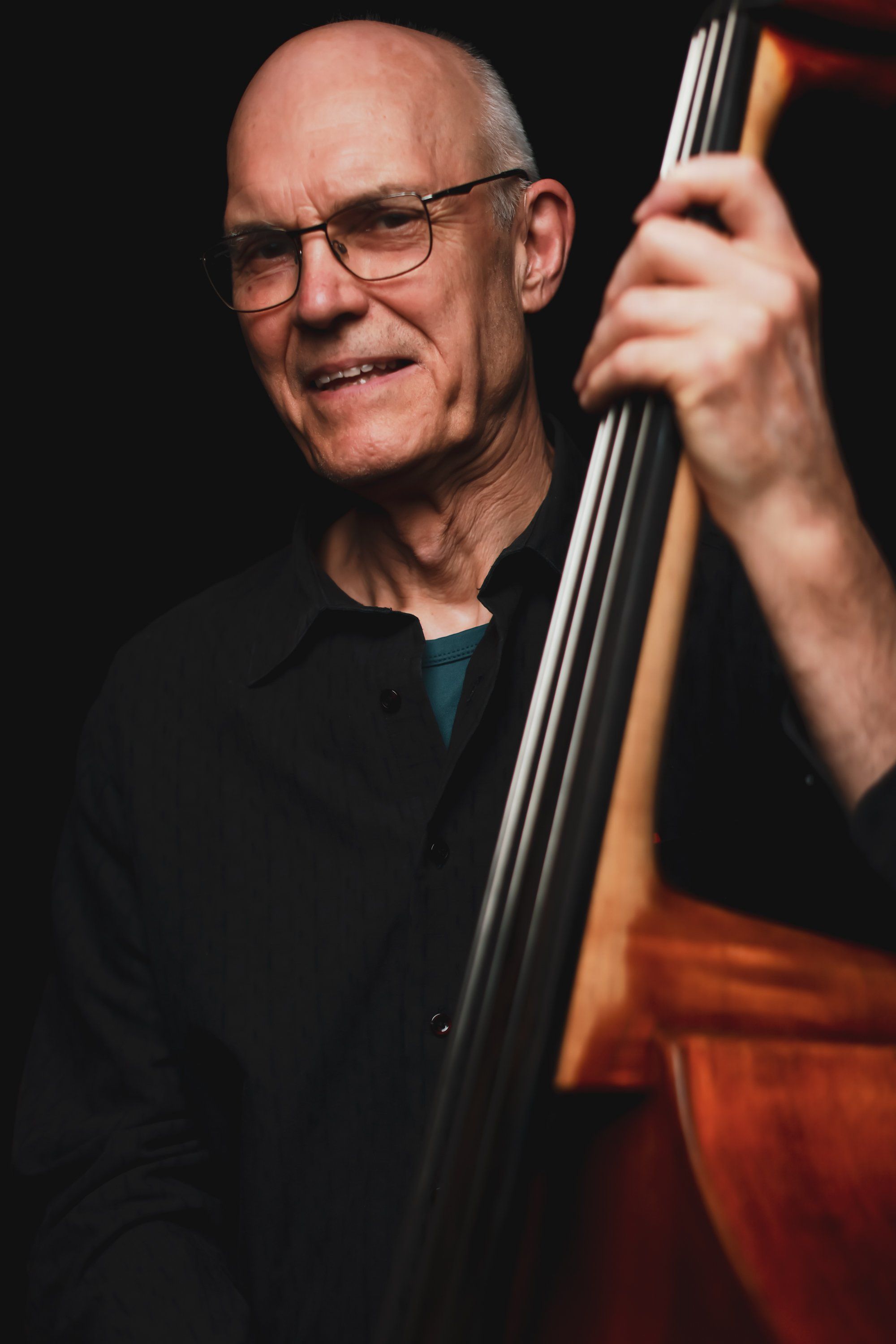 Musician with glasses holding a cello against a dark background, smiling warmly at the camera
