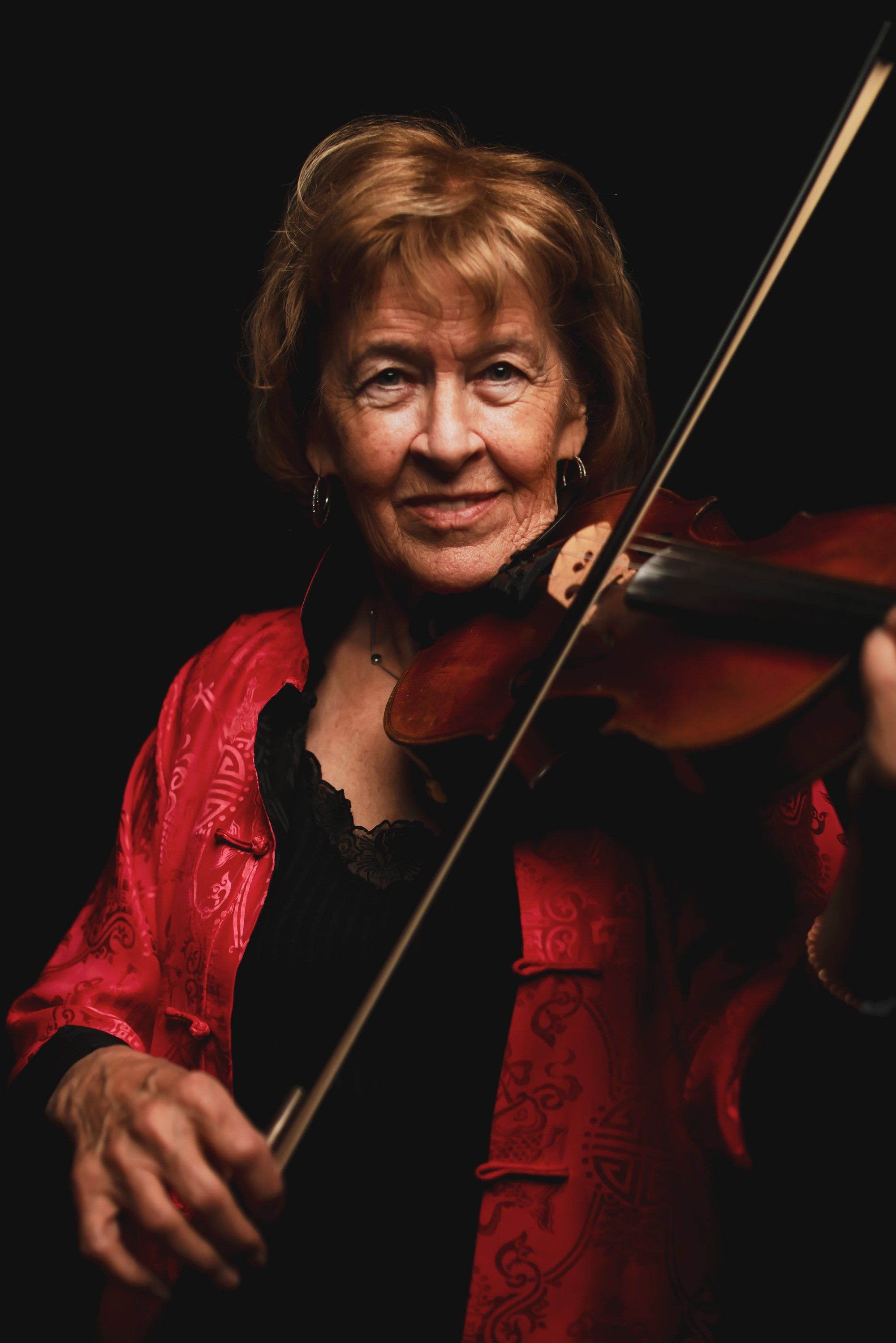 Violinist in red jacket holds bow and instrument against black background, warm side lighting highlighting contemplative expression