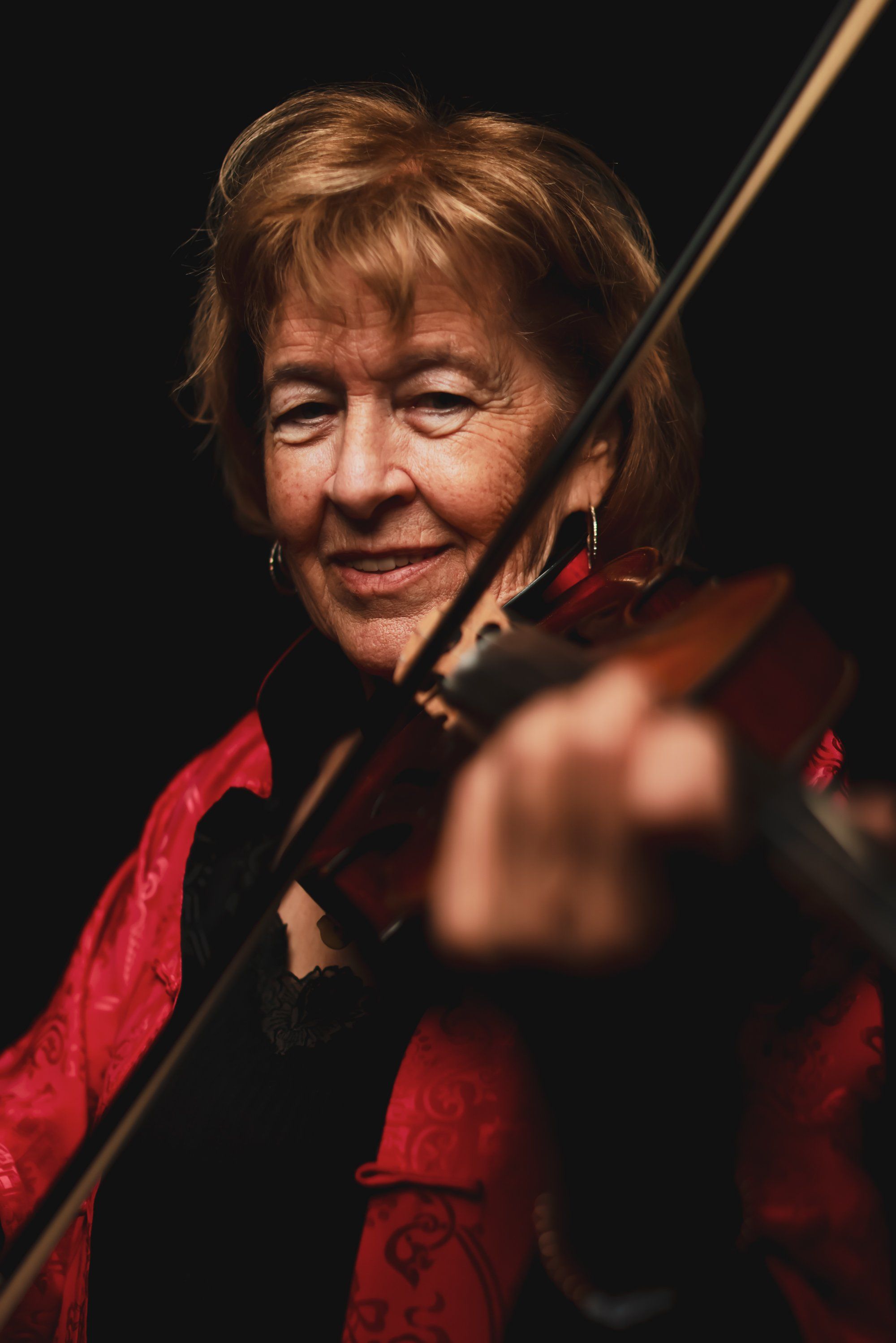 Smiling musician with reddish-brown hair holds a violin against a dark background, wearing a red and black jacket with warm stage lighting