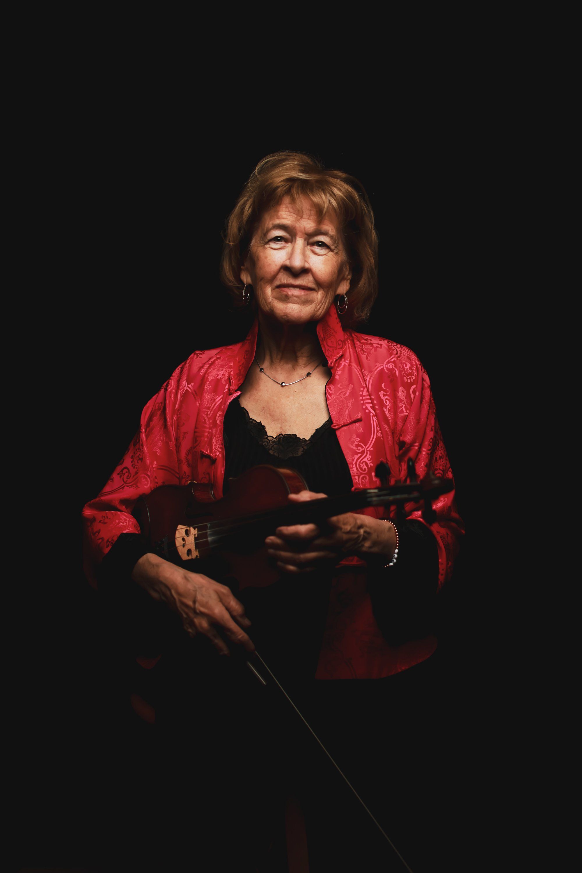 Woman in red jacket holding violin against dark background, warm stage lighting illuminating her face and instrument