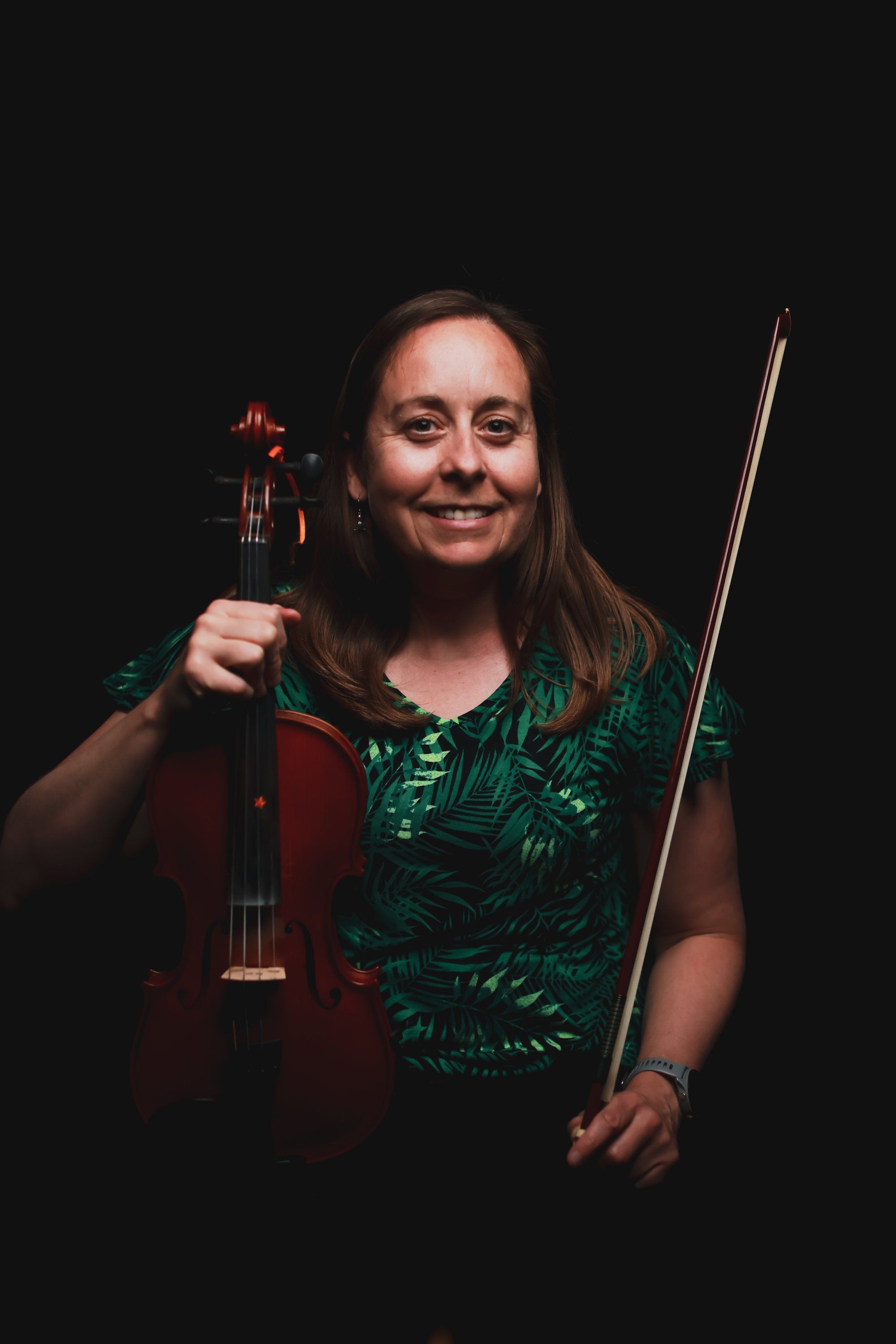 Musician with long dark hair holding a violin and bow against a black background, wearing a green patterned shirt, smiling warmly at the camera