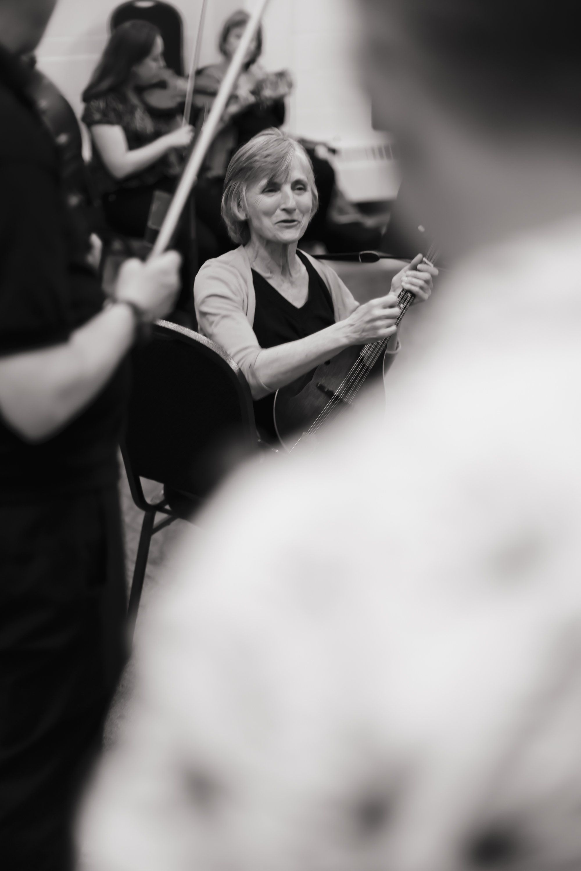 Woman conducting an orchestra with focused expression, surrounded by musicians holding instruments in black and white