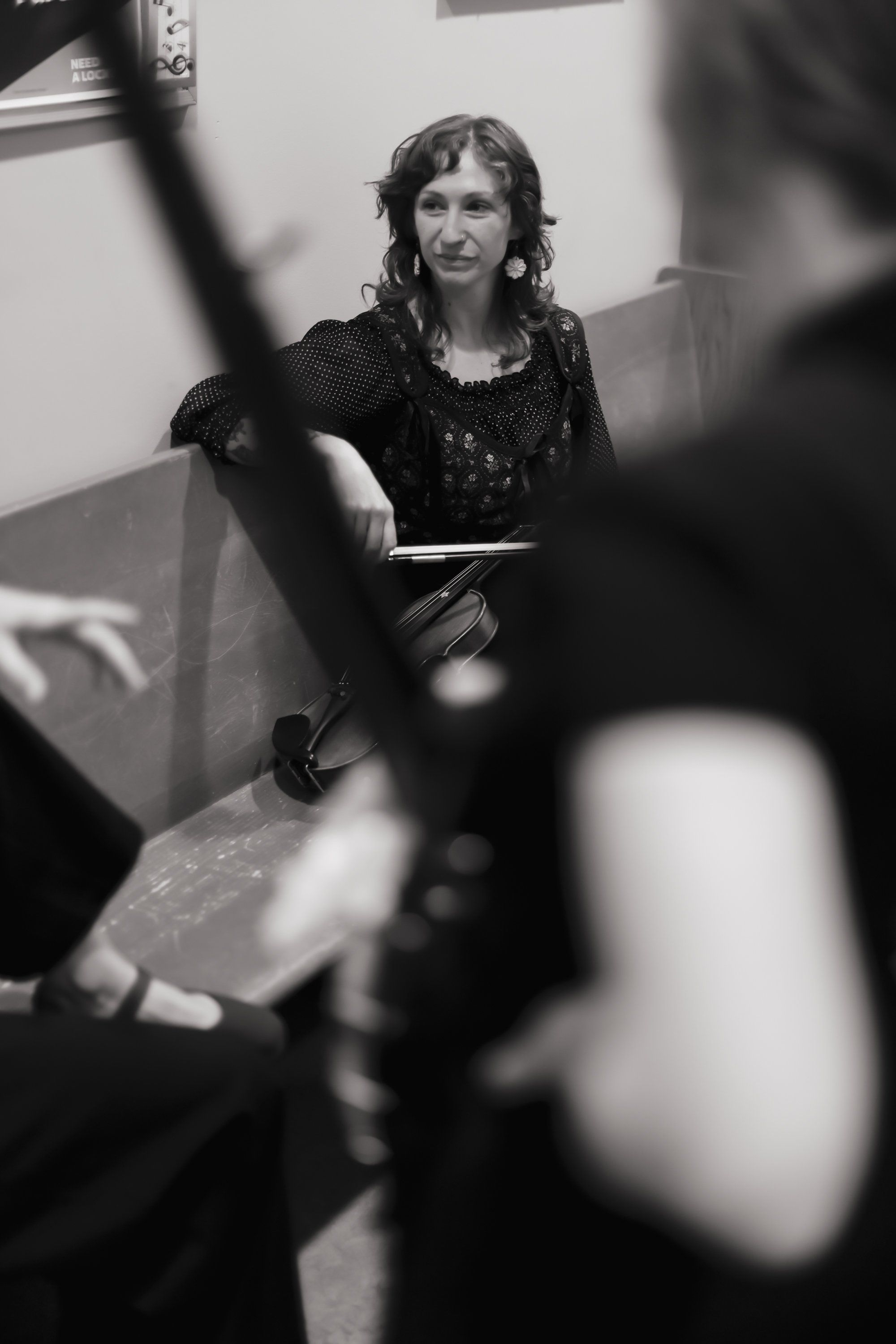 Woman with long dark hair in sequined black dress sits thoughtfully during intimate concert, soft natural light filtering across her face