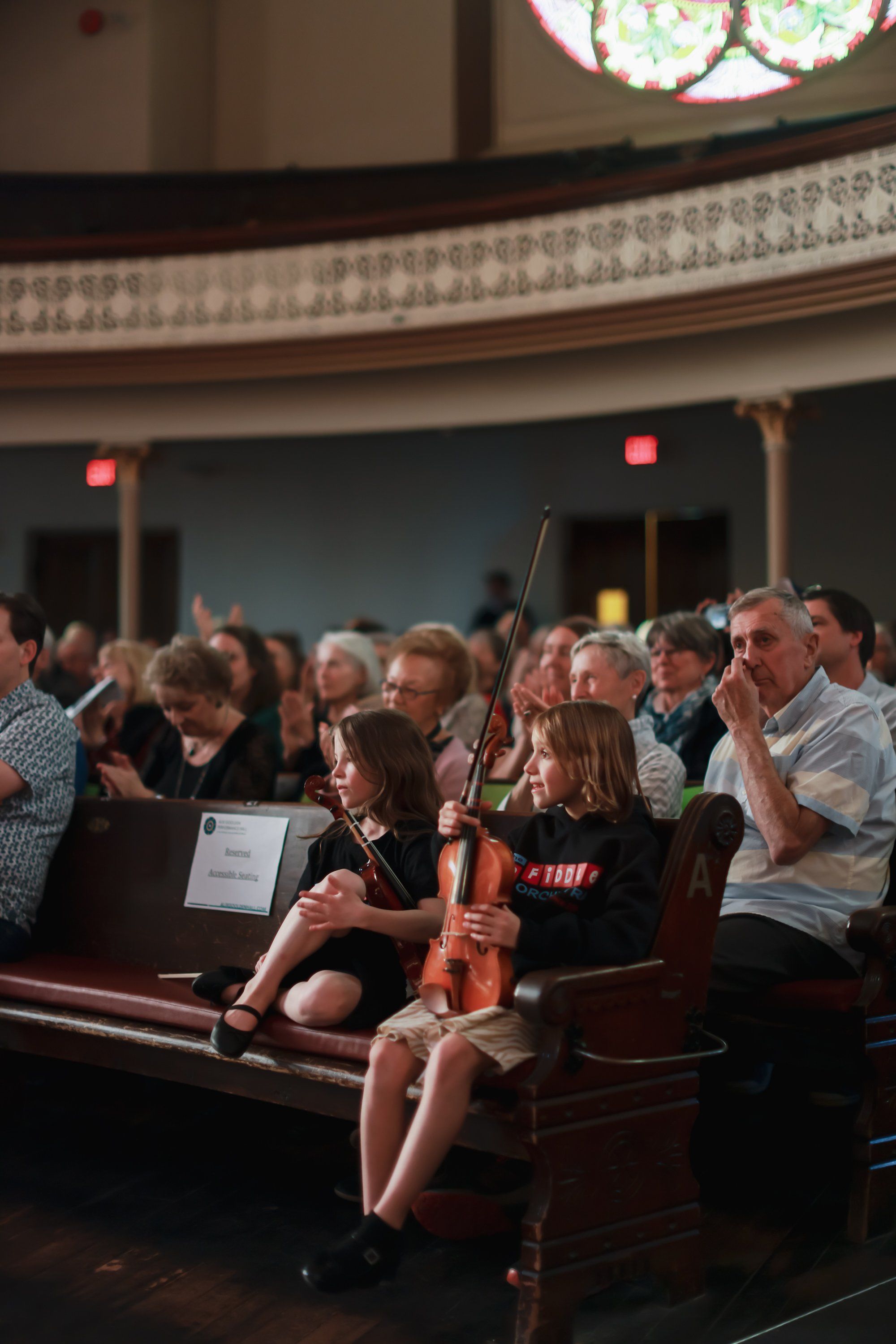 Congregation members stand and applaud during a concert in a wooden church sanctuary, with a young violinist seated in the front pew