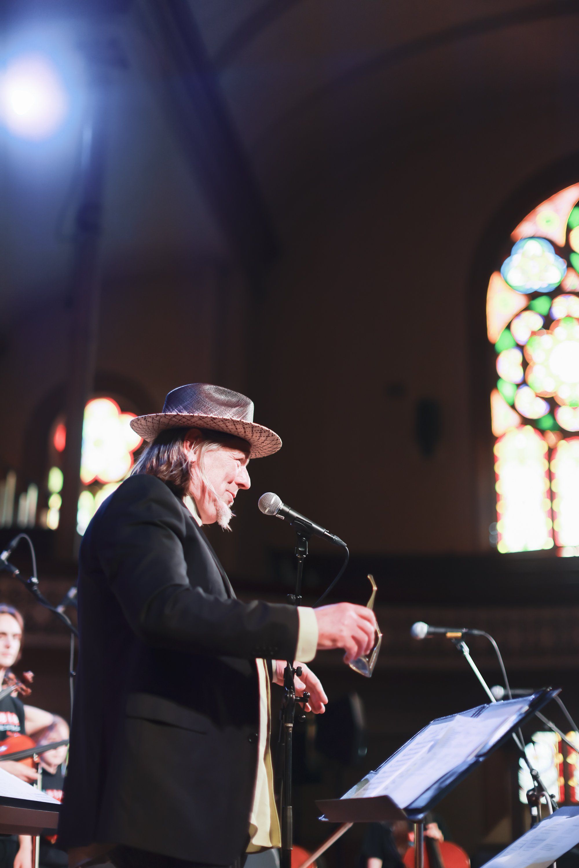 Elderly musician in black suit and cowboy hat performs at microphone in dimly lit venue with colorful stained glass window glowing behind him