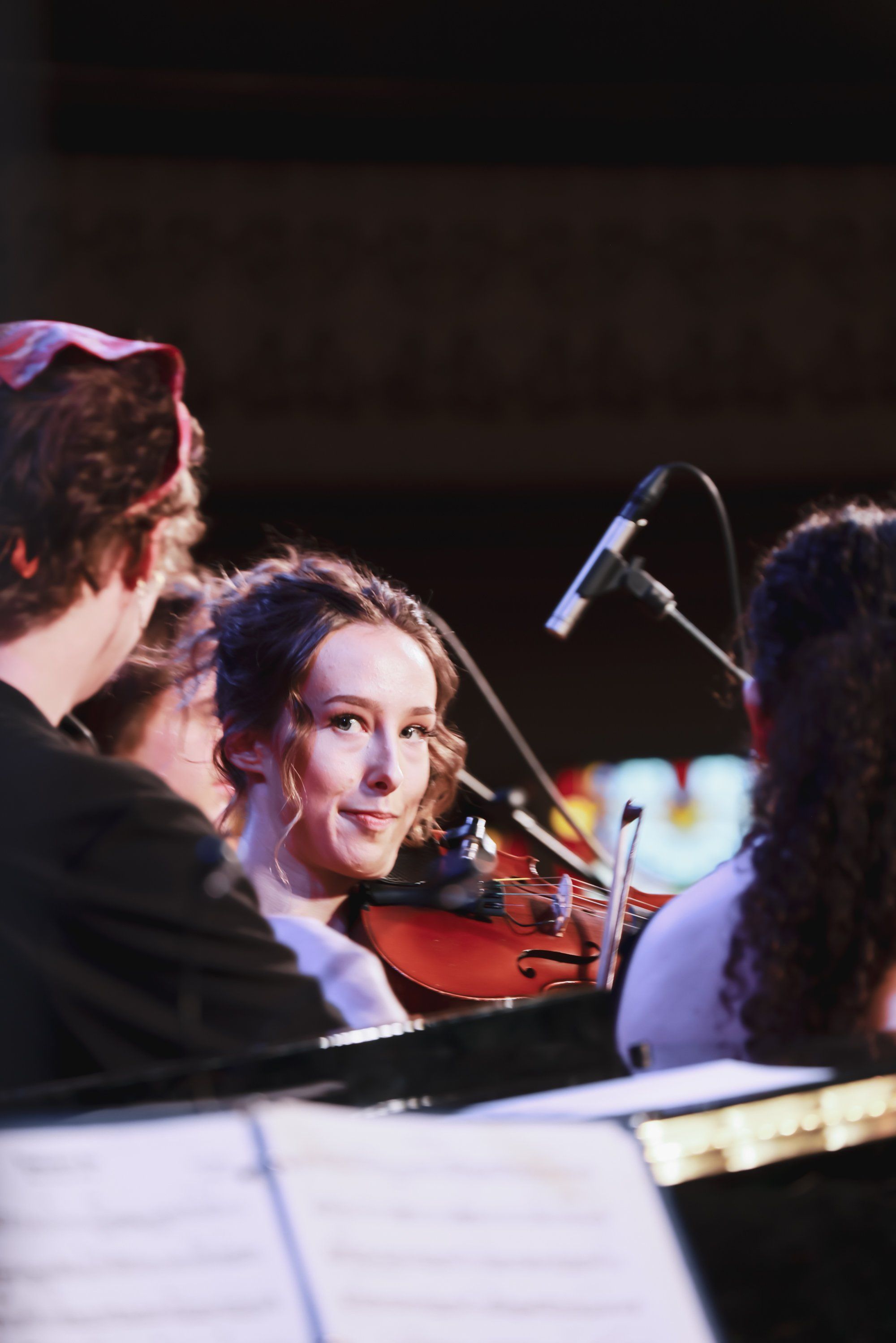 Violinist in concert lighting performs beside sheet music and microphone, surrounded by ensemble members in warm stage glow