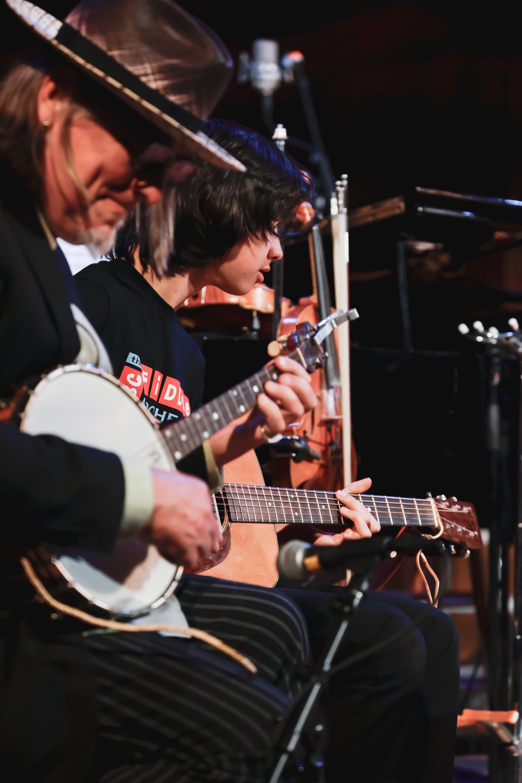 Two musicians perform on stage under warm lighting, one wearing a cowboy hat playing banjo while the other plays acoustic guitar, both focused intently on th…