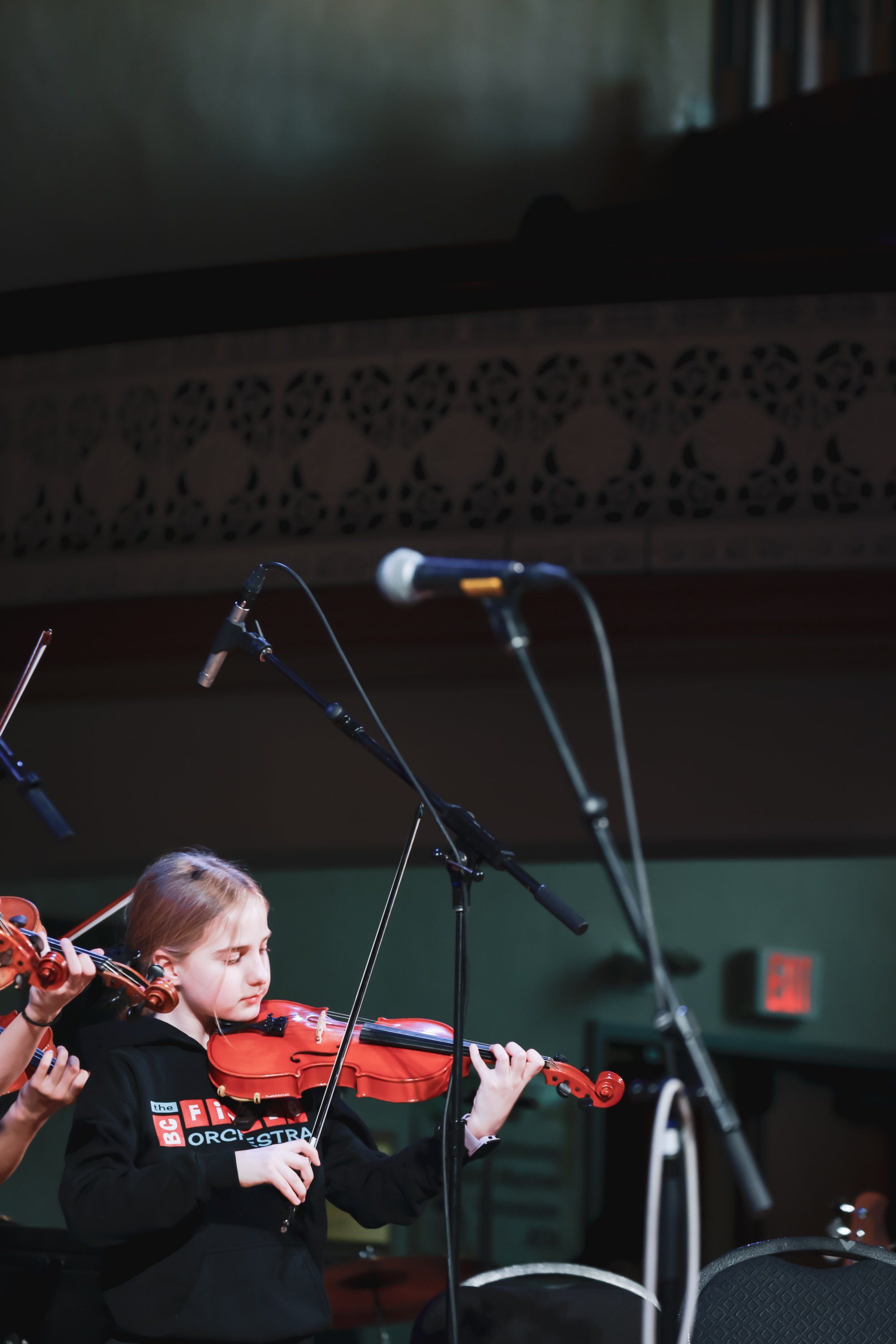 Young violinist performs under stage lights, bow drawn across instrument in concentrated focus, microphone positioned nearby on darkened concert stage