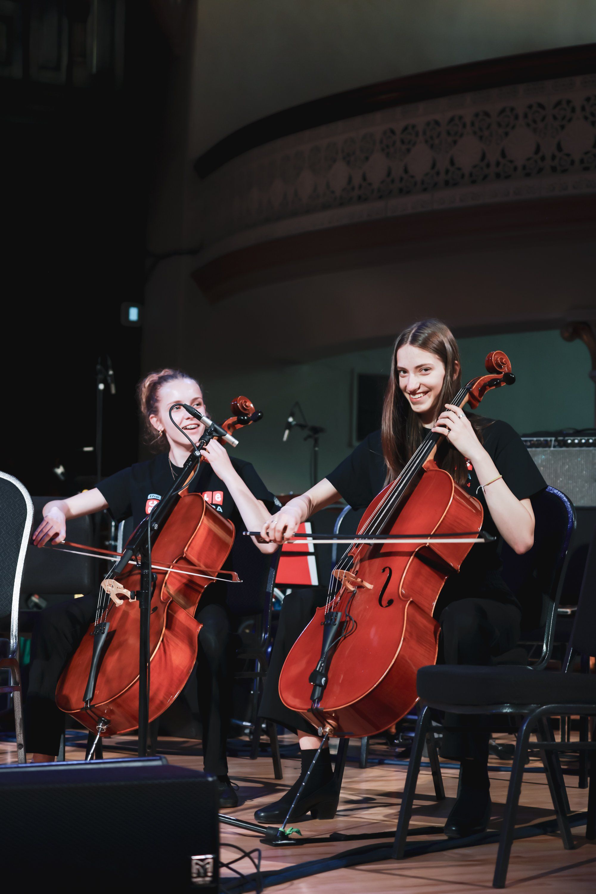 Two cellists perform on stage under warm spotlights, their instruments glowing amber against the dark auditorium backdrop