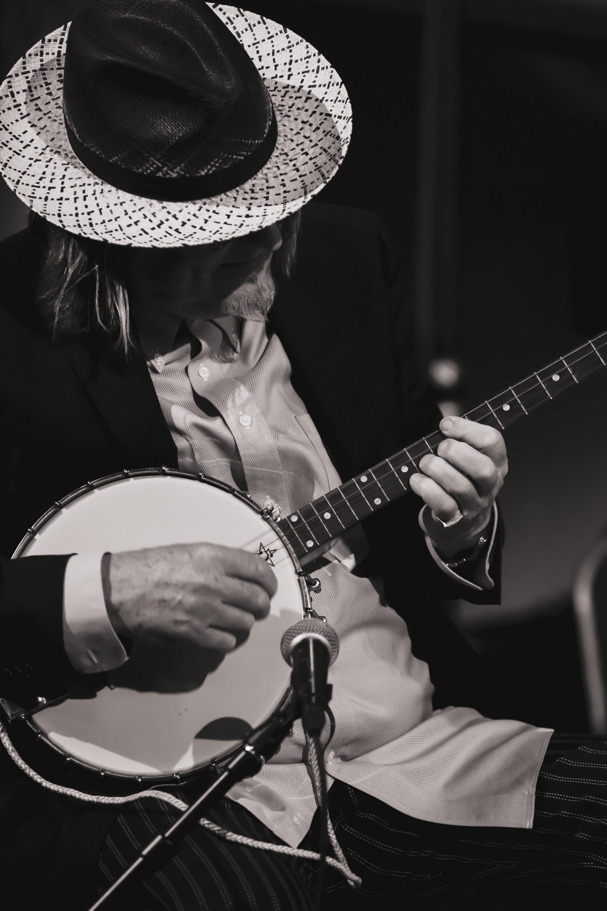 Musician in wide-brimmed hat playing banjo at microphone, captured in dramatic black and white against dark background