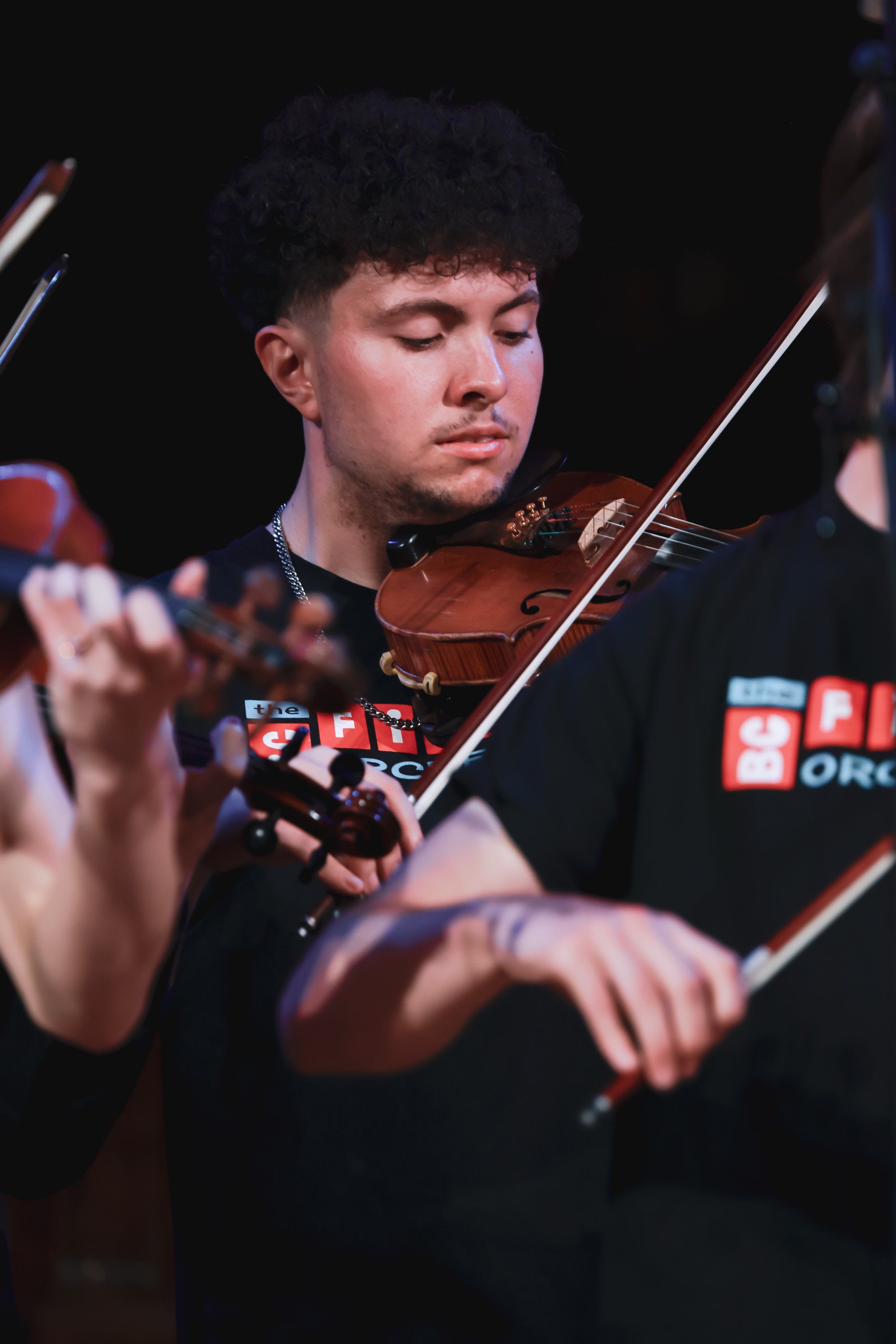 Young violinist in black performance attire concentrates intently while playing, warm stage lighting highlighting the instrument and focused expression
