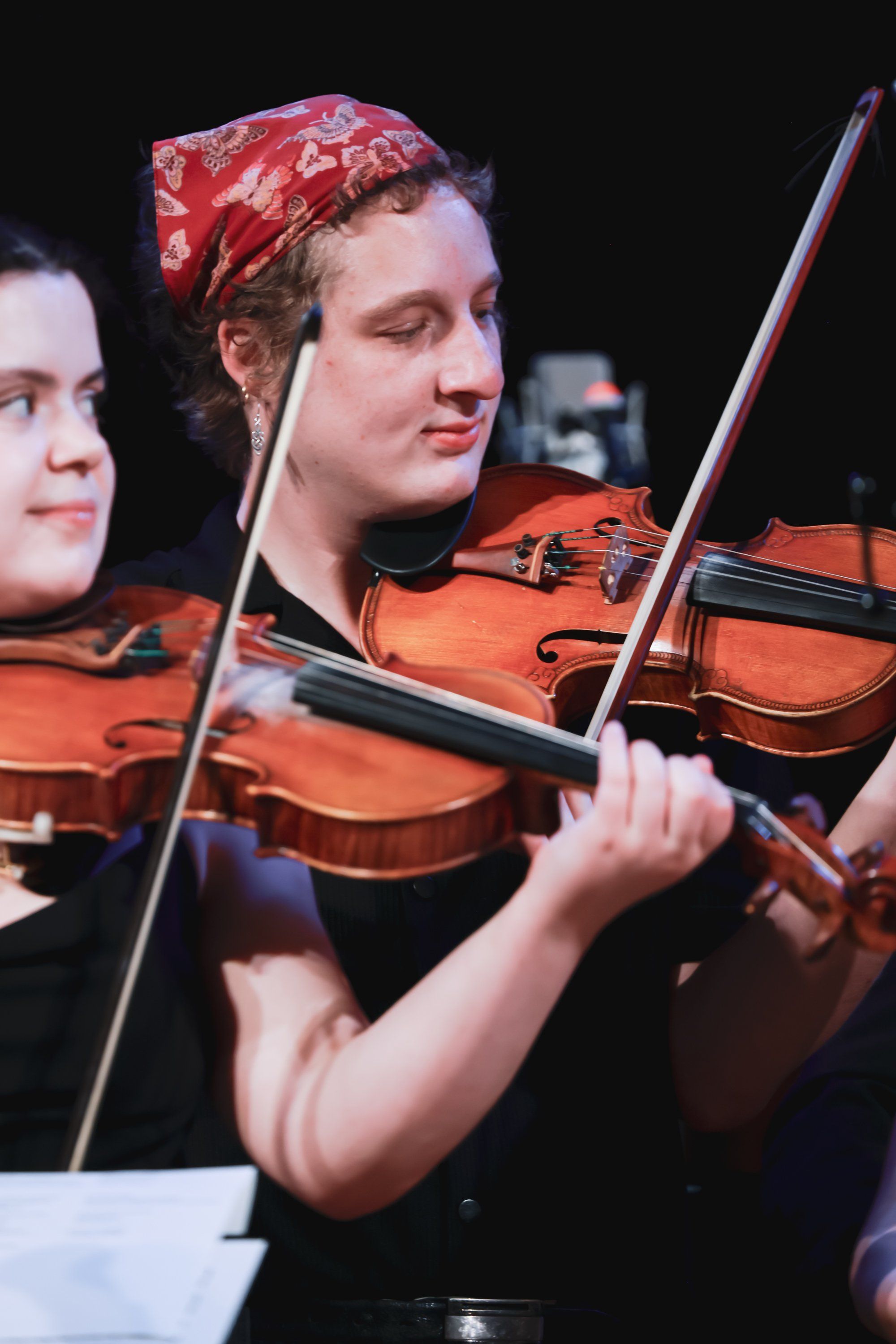 Violinist in red patterned headwrap performs with focused expression during ensemble concert against dark background