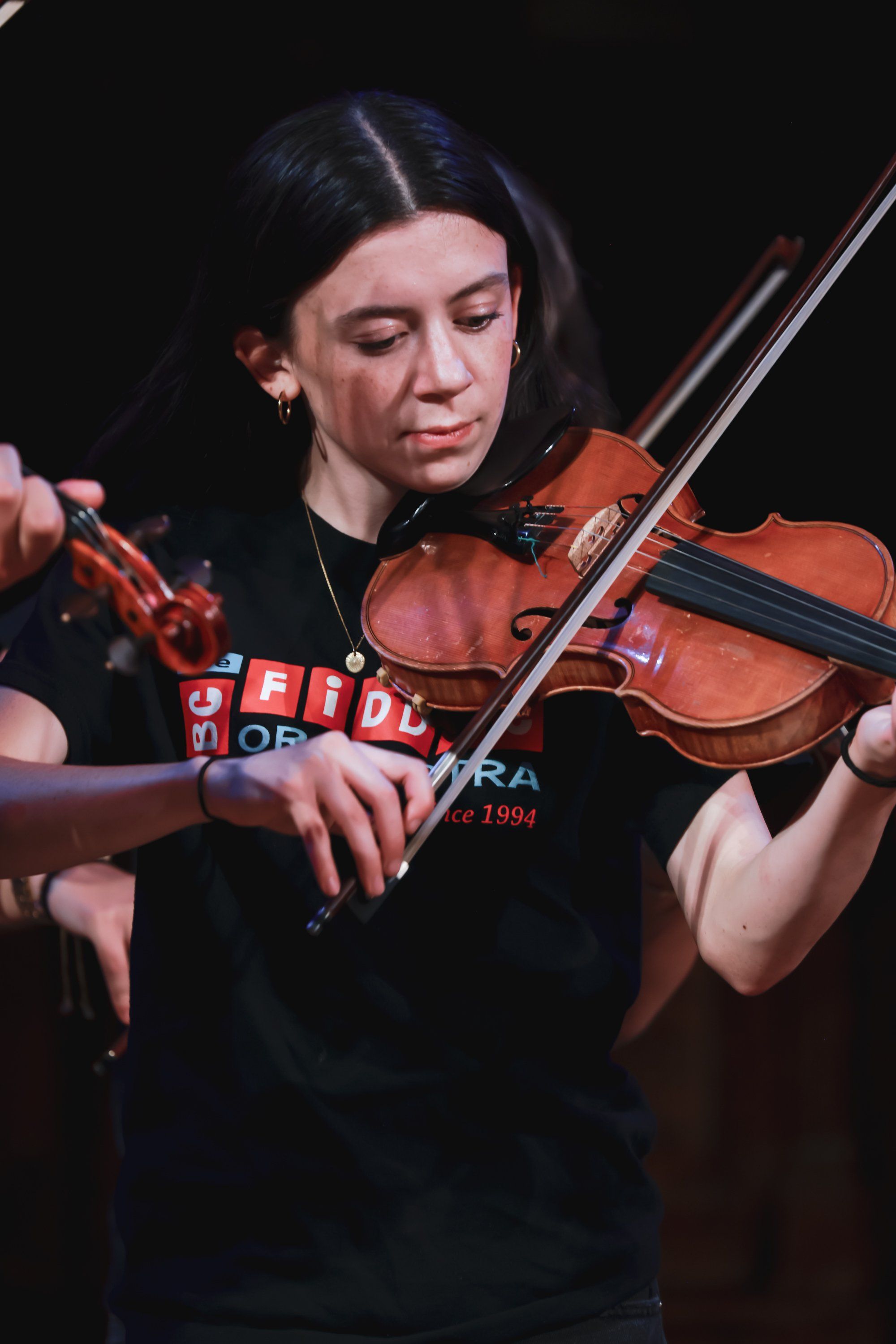Violinist in black performing intently against a dark stage, instrument held at playing position with focused expression