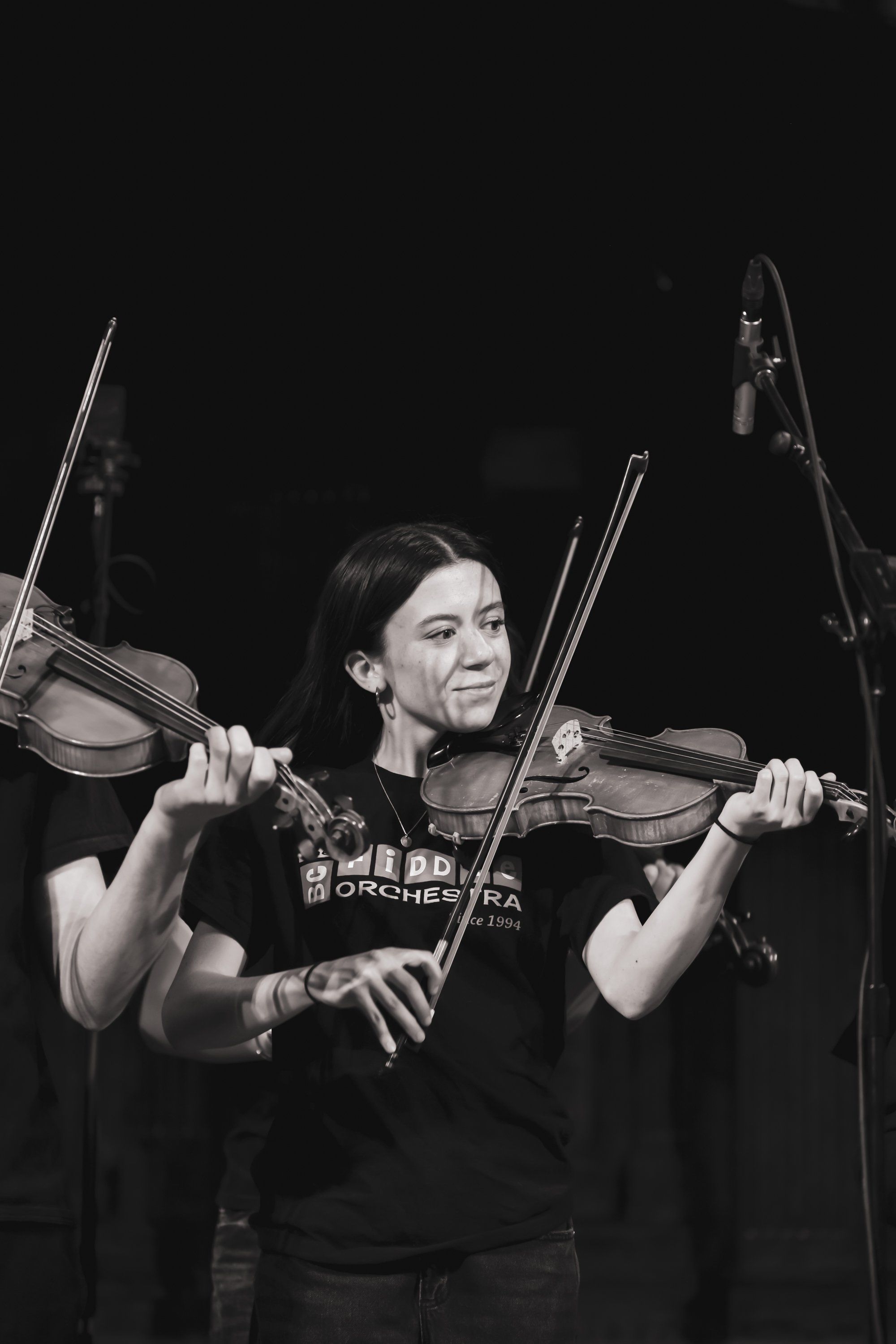 Violinist in black concert tee performs against dark background, holding dual instruments with focused expression and dramatic side lighting