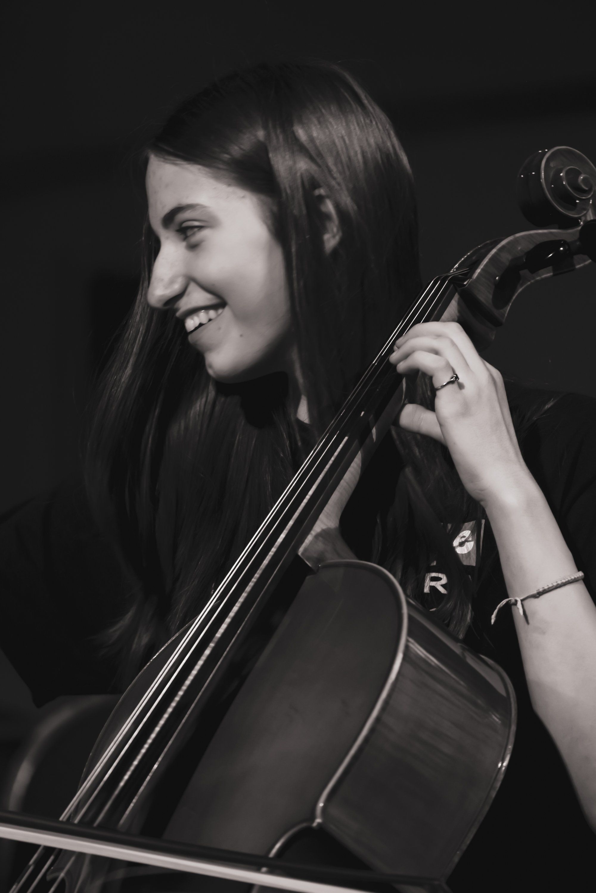 Smiling cellist with long dark hair holds her instrument against a dark background, captured in profile during an intimate performance moment